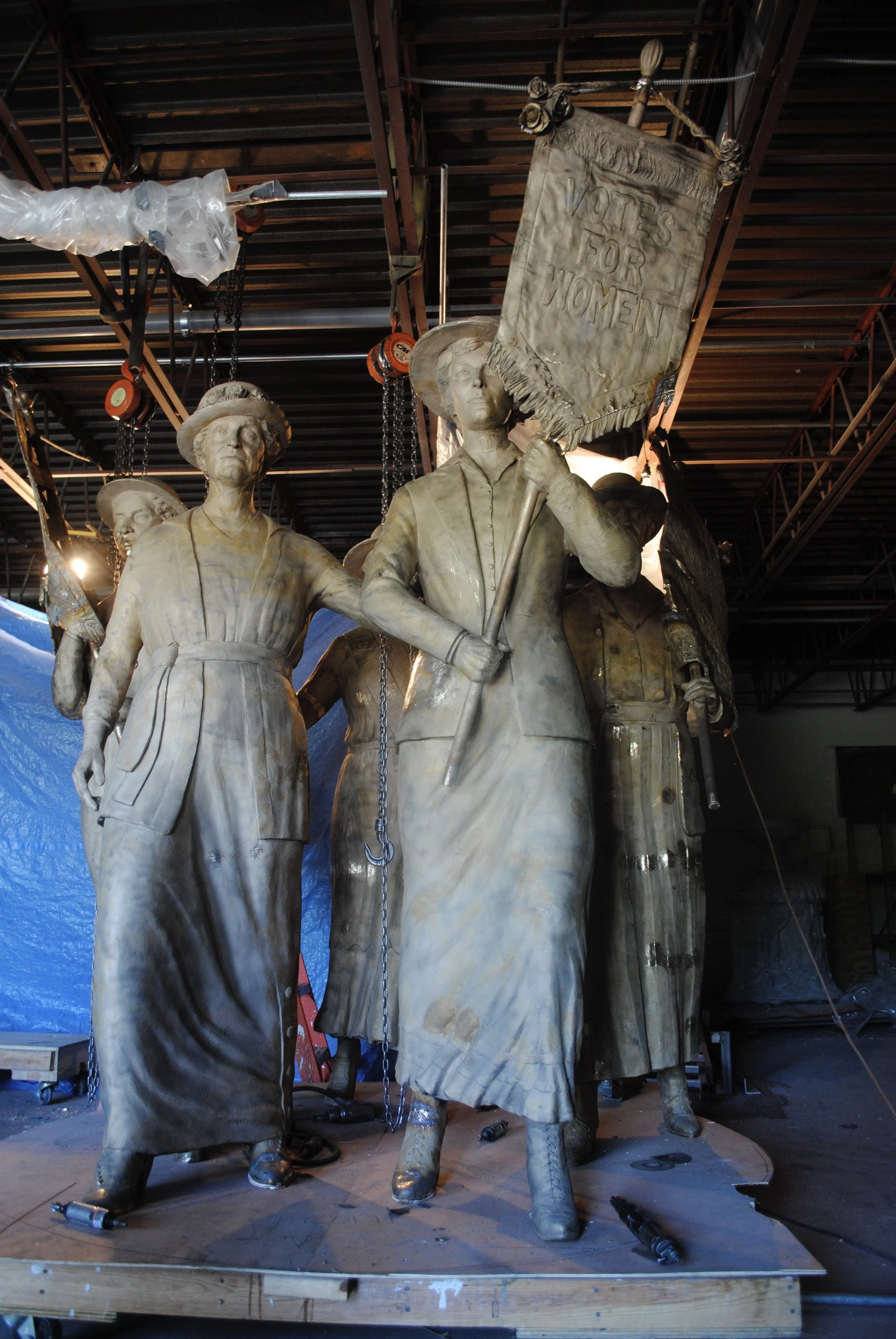 A group of large, unfinished bronze statues of women, one holding a sign that reads 'Votes for Women,' in a foundry. The statues are surrounded by tools and scaffolding.