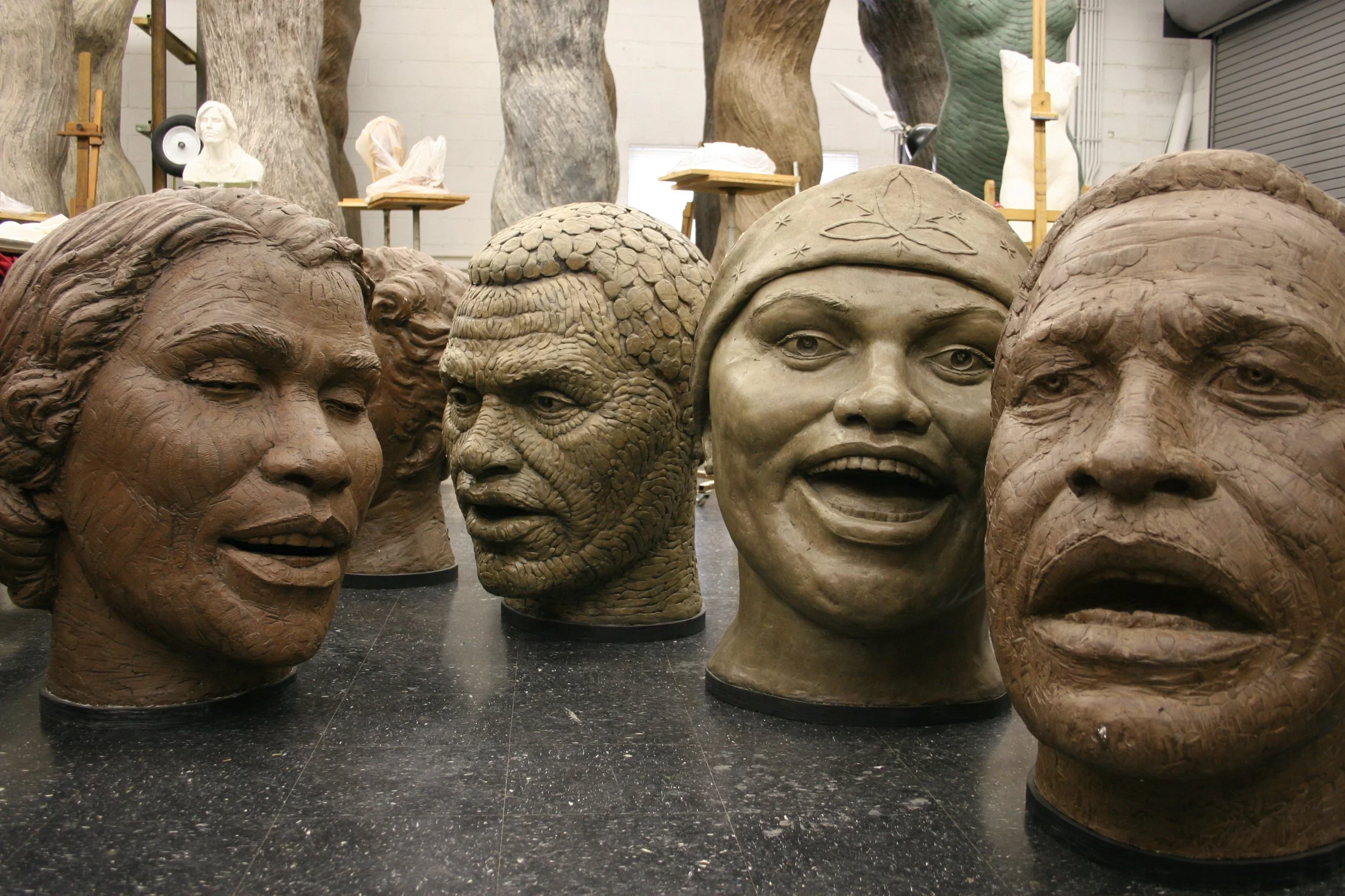 Multiple large sculpted busts of diverse faces arranged in a studio setting, with some sculptures in progress and busts displayed on stands in the background.  Cultural Heroes