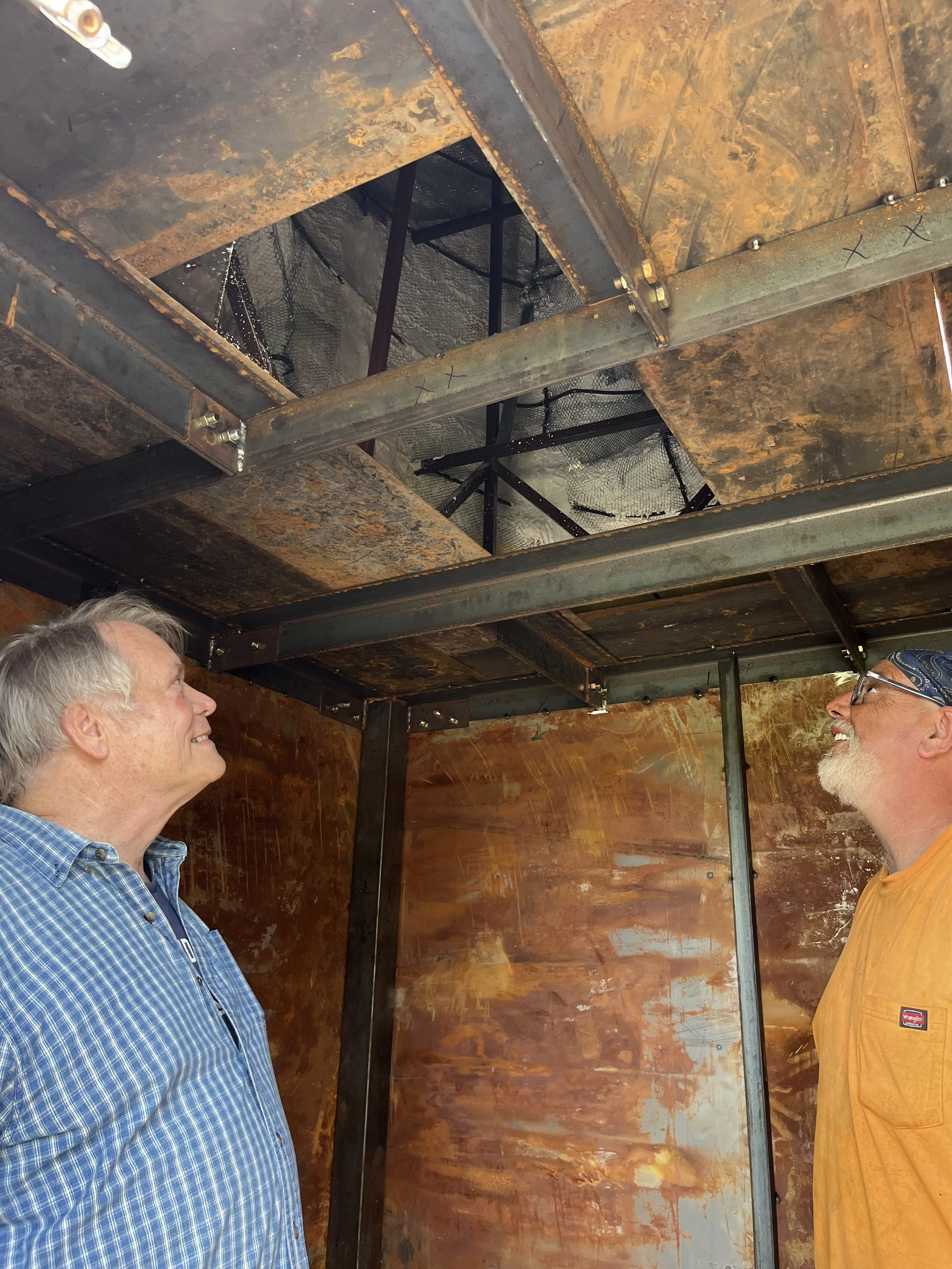 Two men standing in a room with wooden walls, facing each other and smiling. The ceiling has a rusted metal frame and some grid-like metal structures.
