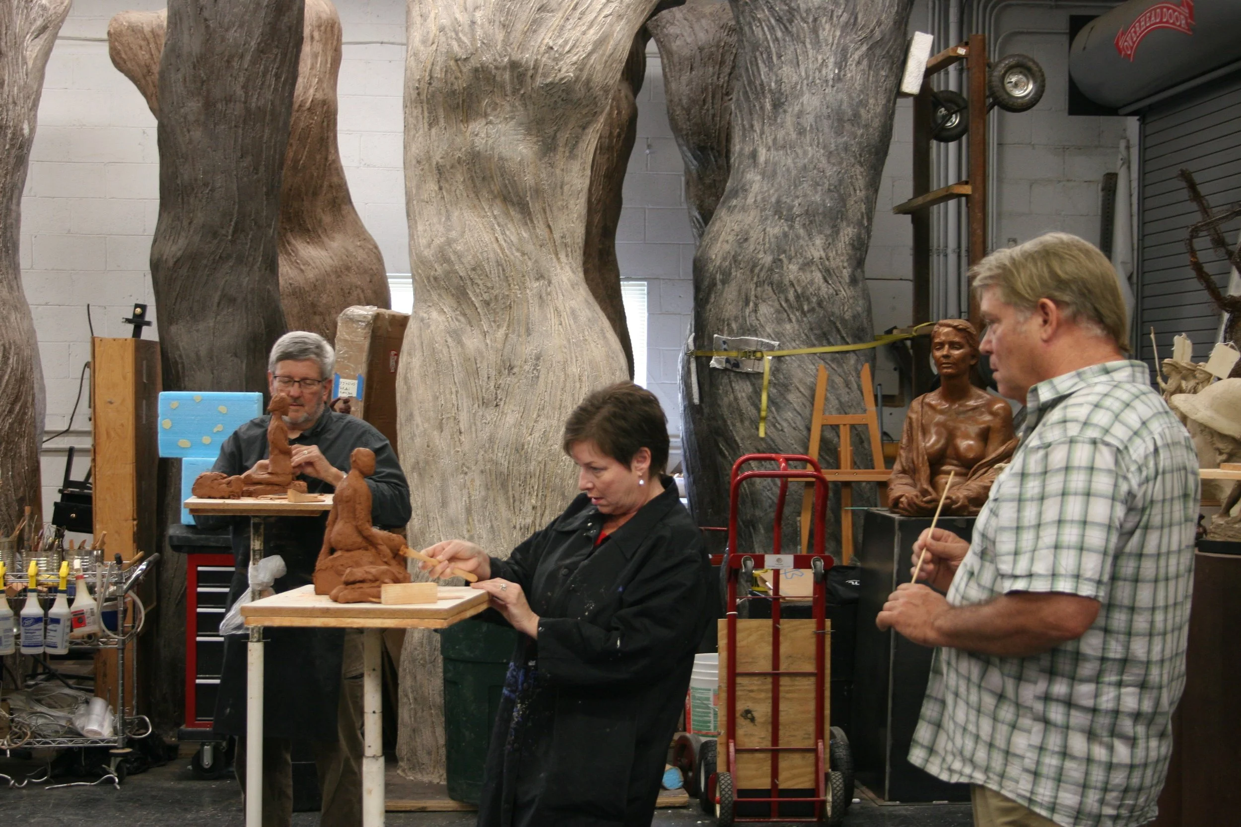 People working on clay sculptures in an art studio with large wood-textured sculptures nearby.