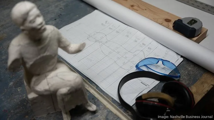 A workspace with a small sculpture of a person sitting, blue safety glasses, a tape measure, a large sheet of grid paper with a diagram, and a ruler on a workbench.