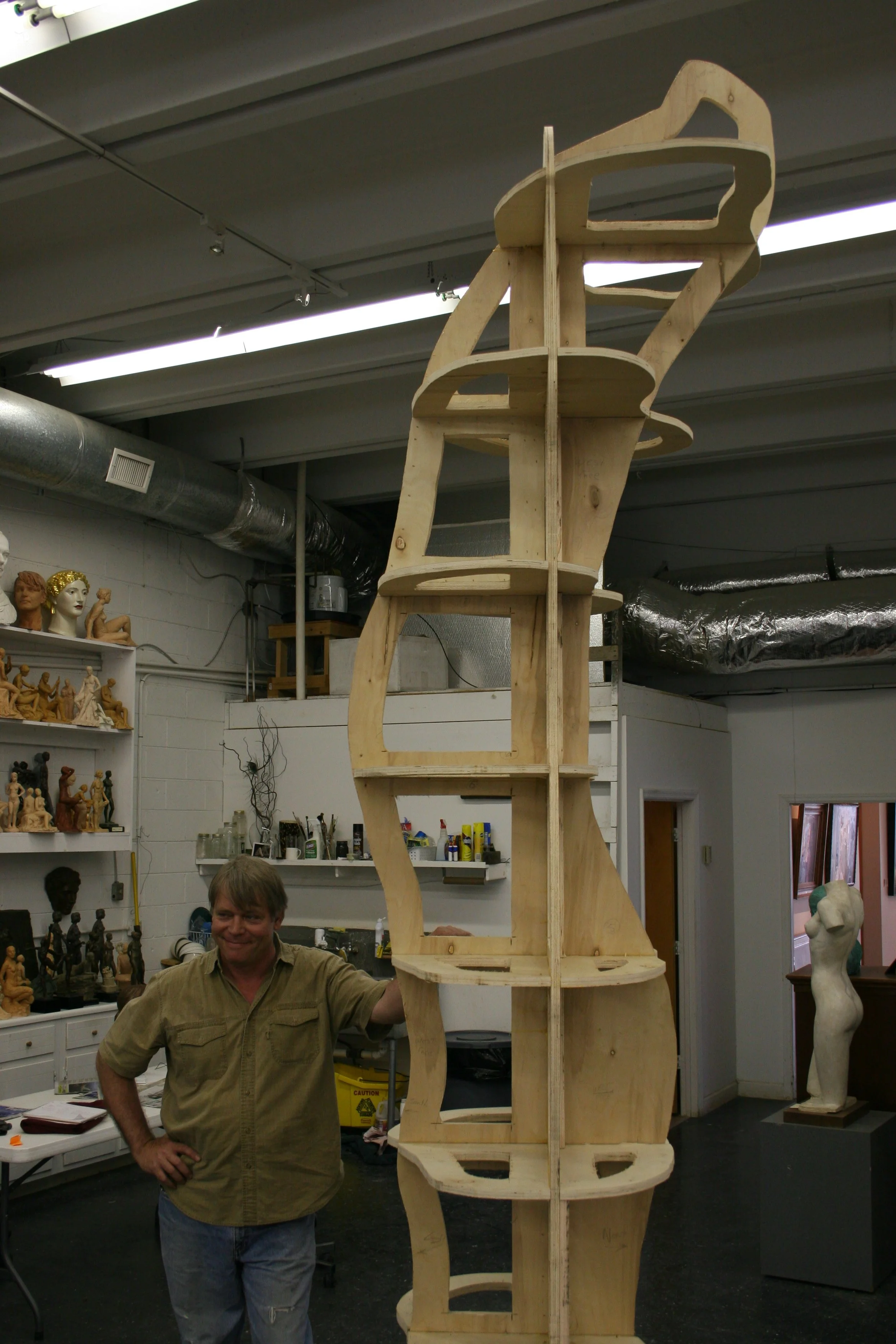 Artist Alan Lequire standing in a workshop next to a tall, multi-level interior armature structure with curved and open sections. Shelves and bust sculptures are visible in the background.