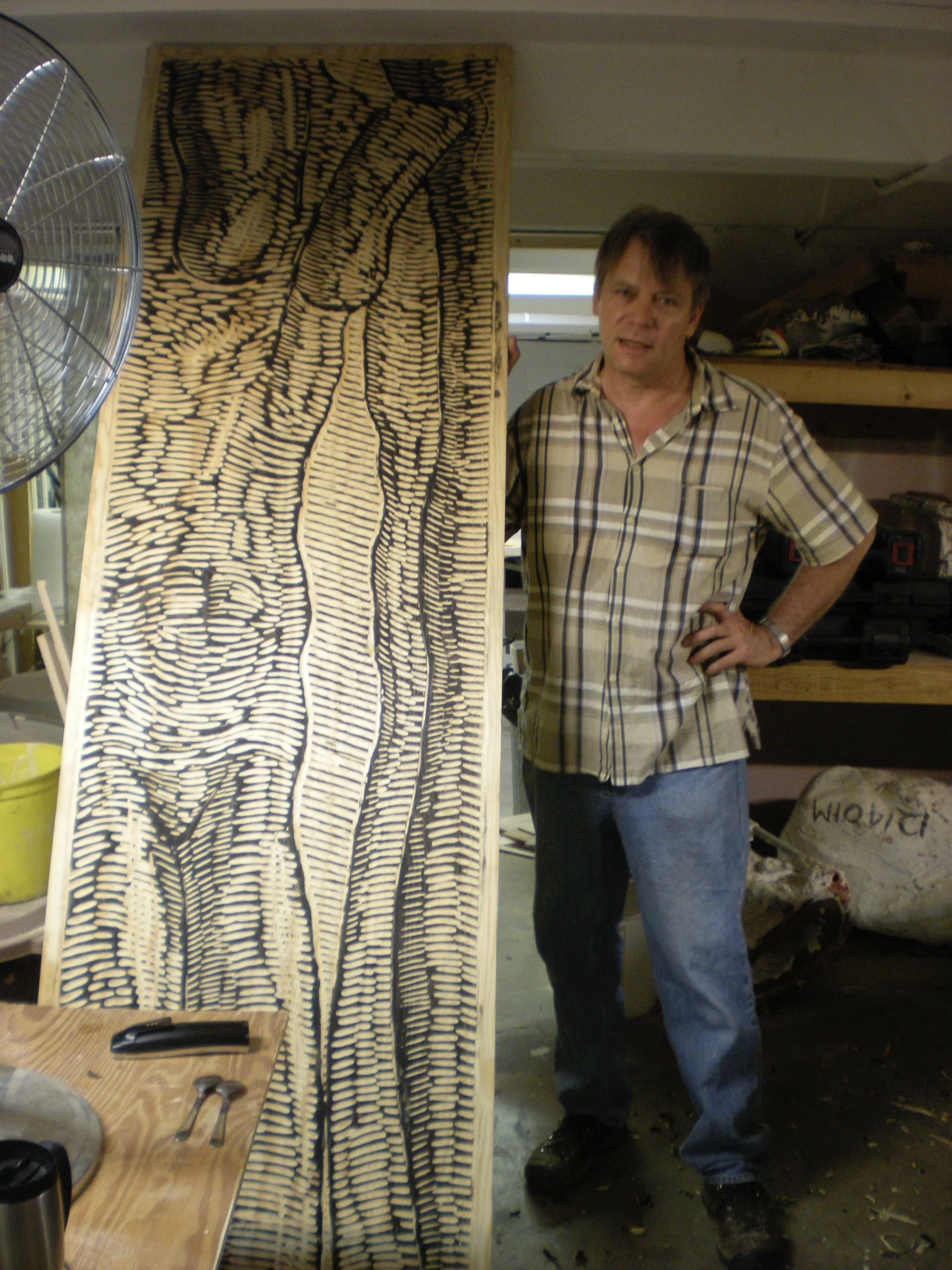 A man stands in a workshop next to a large wood block print featuring a detailed black and beige design of a hand holding a bone. The workshop has tools on a workbench, a fan, and storage shelves.