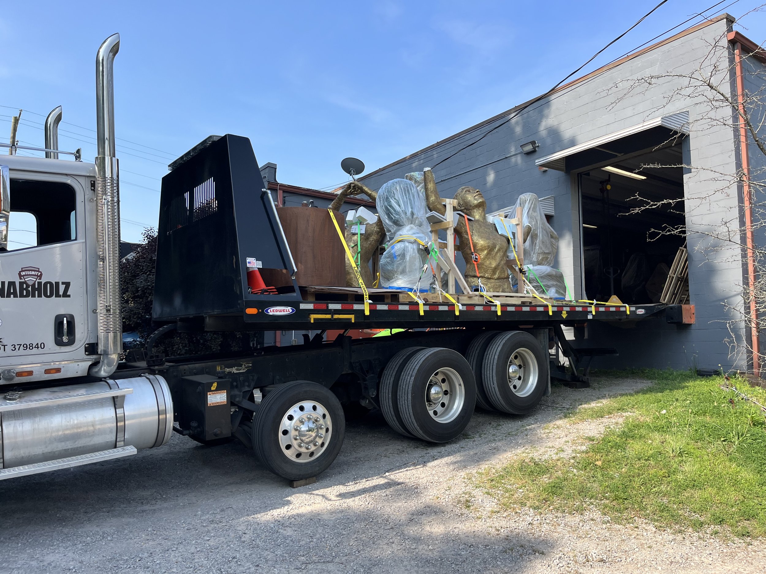 A flatbed truck carrying two large golden statues of women, with one woman raising her arms and the other with her hands on her head, in front of a gray building with an open garage door and a clear blue sky.