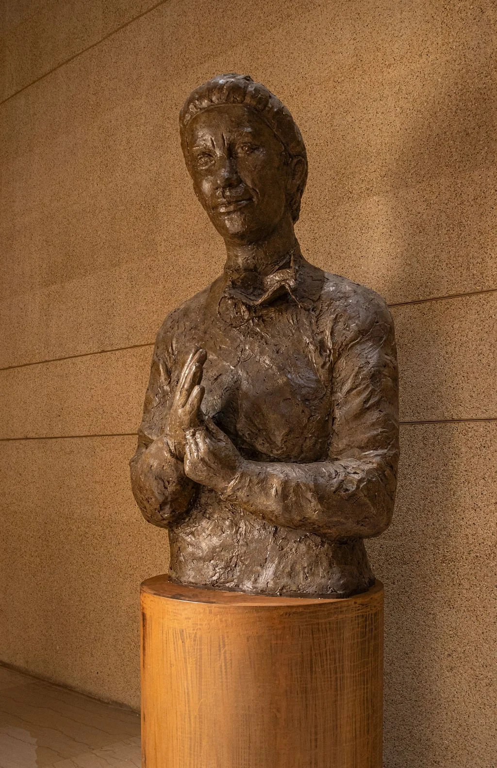 Bronze sculpture of a woman with short hair, holding a book in one hand and the other hand placed over her chest, displayed on a cylindrical metal pedestal.