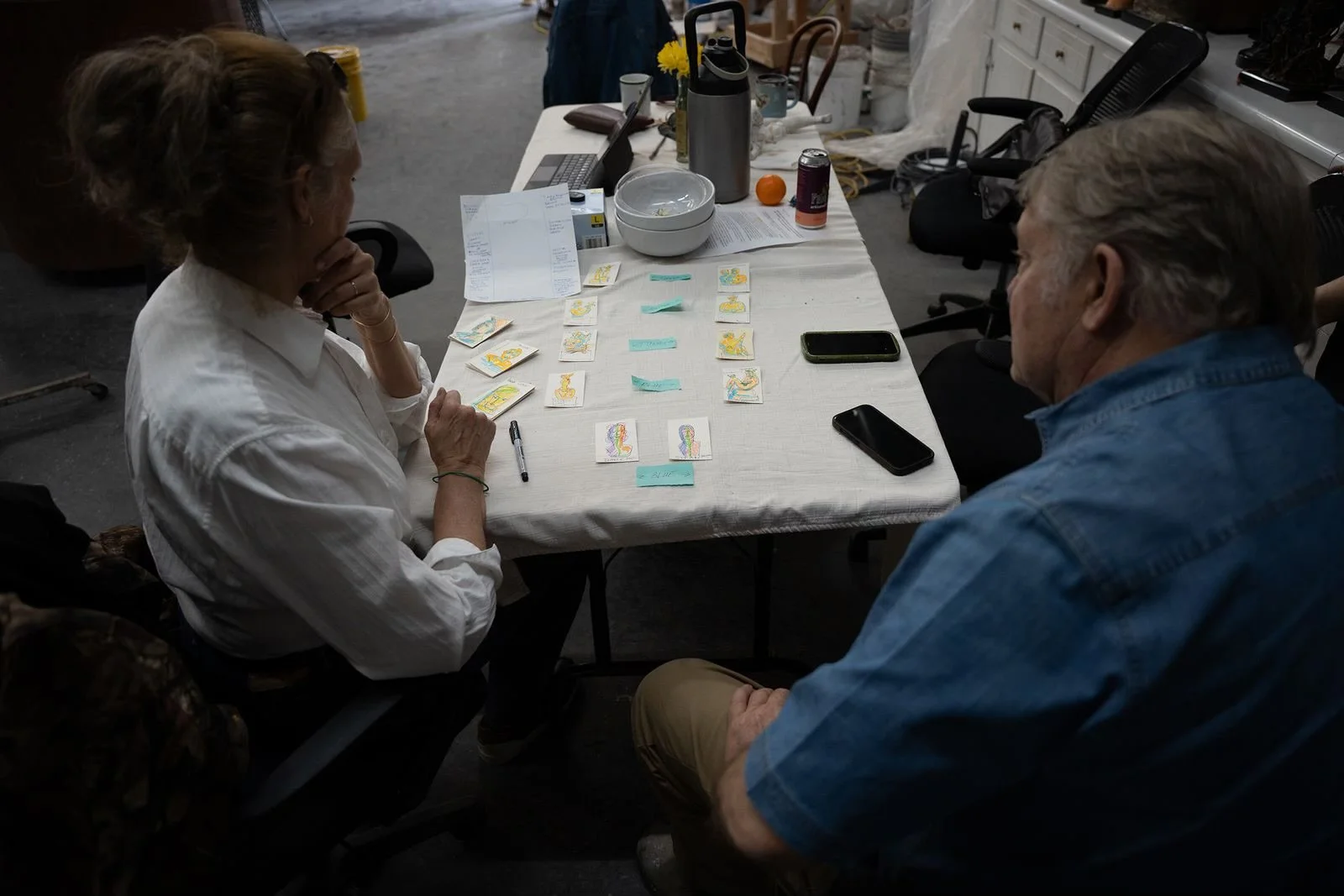 People sitting at a table playing a card game with colorful illustrated cards, surrounded by various items including phones, notebooks, and drinks.