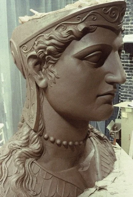A detailed sculpture bust of a woman with intricate hair, a headpiece, a beaded necklace, and a serious expression, set against a workshop background. Athena Parthenos in Parthenon Museum Nashville. 