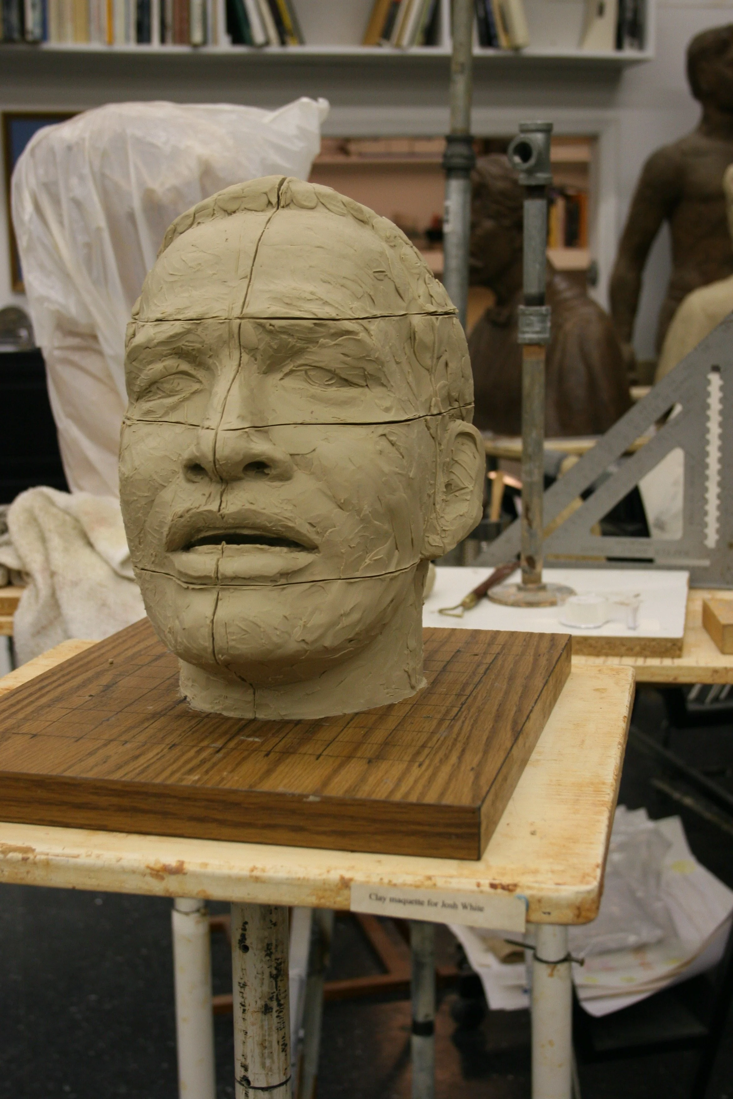 A clay sculpture of a human head on a wooden base, with detailed facial features and markings. The sculpture is in an artist's studio surrounded by sculptures and art supplies.