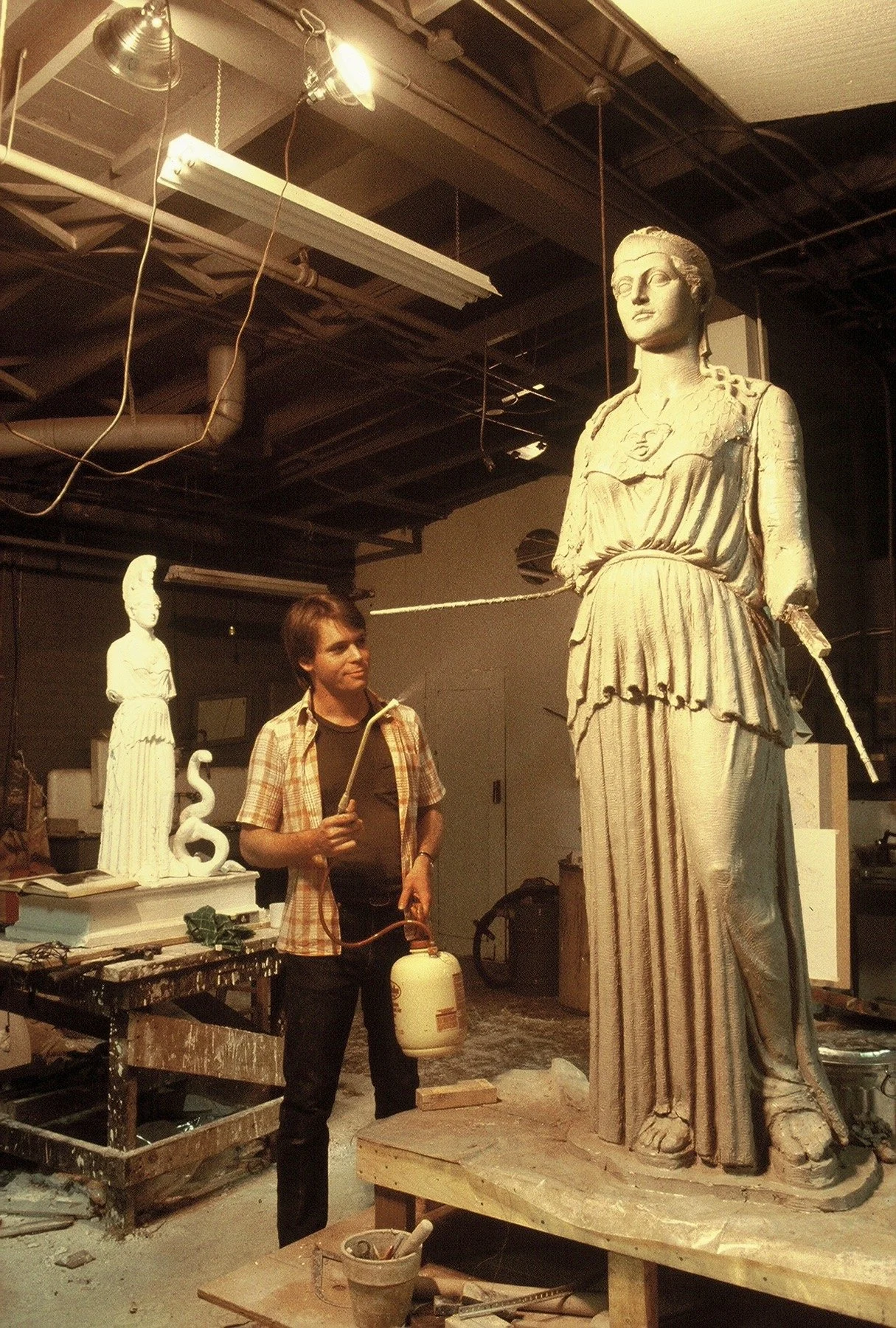 An artist working on restoring a large statue of a woman in an industrial studio. The woman is dressed in ancient attire. Athena Parthenos in Parthenon Museum Nashville. 