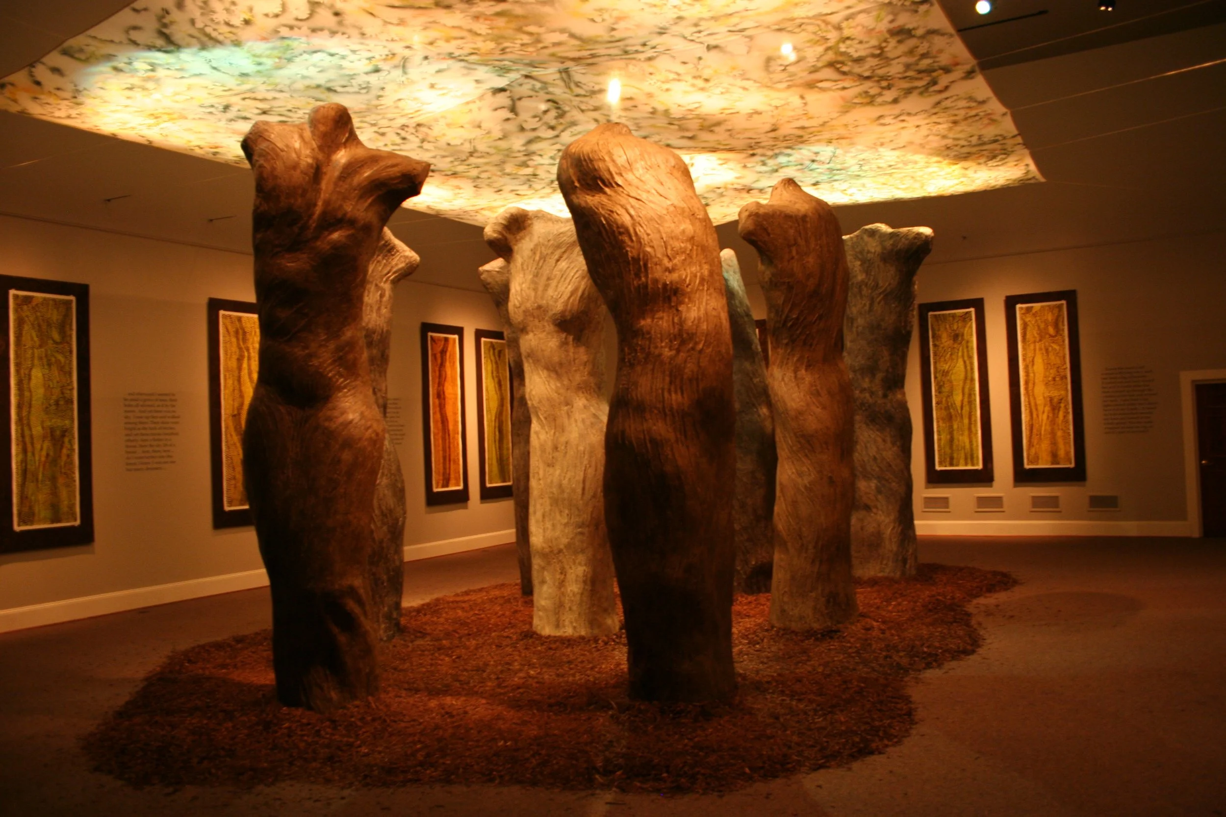 An art installation of tall, sculpted wooden logs with a colorful, illuminated ceiling above them, displayed in a gallery setting. Dream Forest Installation.