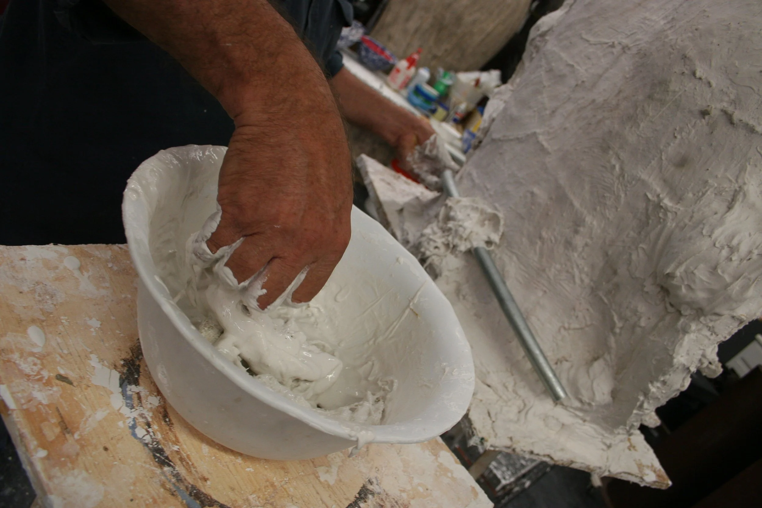 Person mixing white plaster in a bucket at an art studio, with tools and supplies in the background.