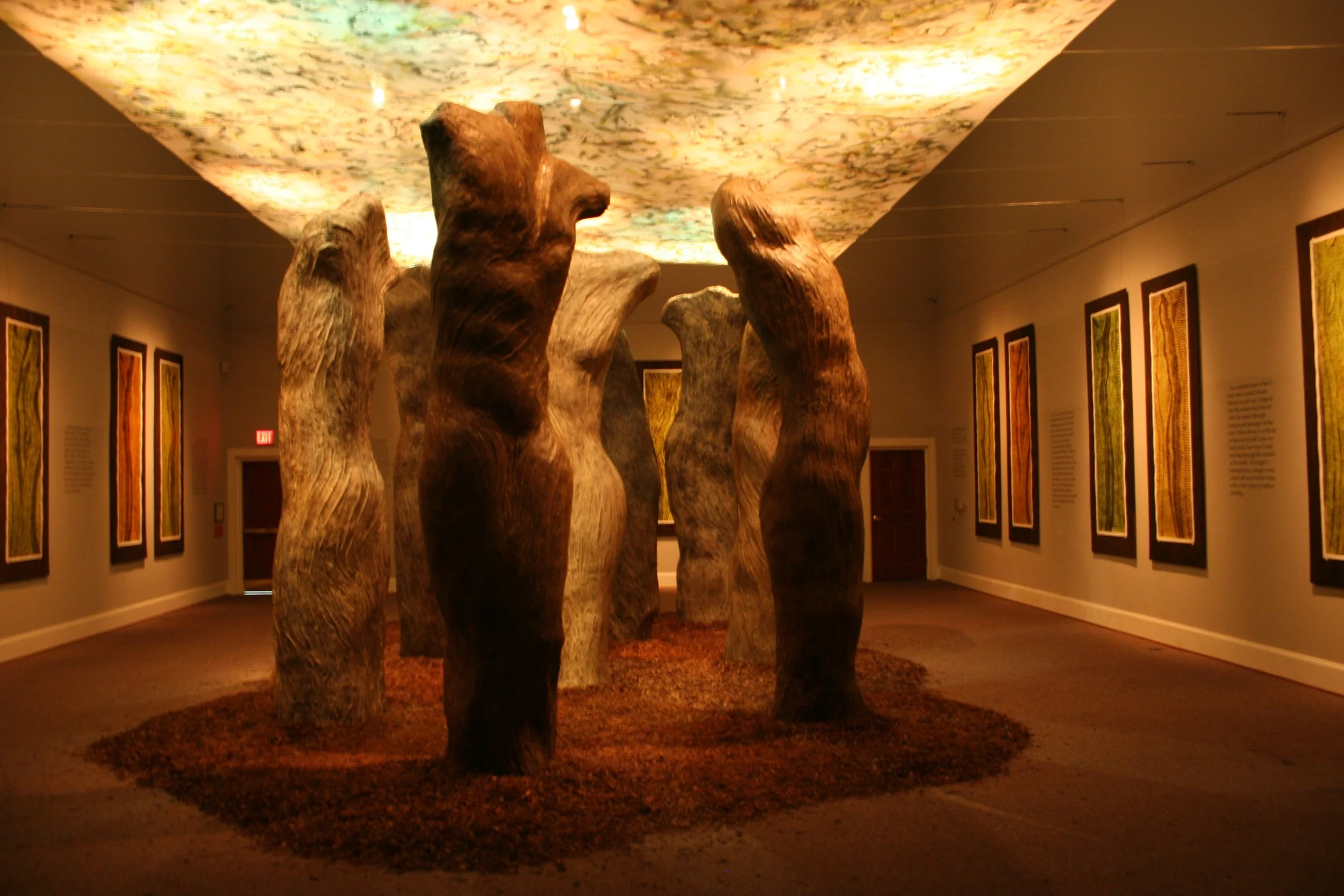 An art installation featuring large, abstract, wooden sculptural figures arranged on a floor covered with reddish-brown material. The background shows framed artworks on the wall and an illuminated ceiling with a textured, multicolored pattern. Dream