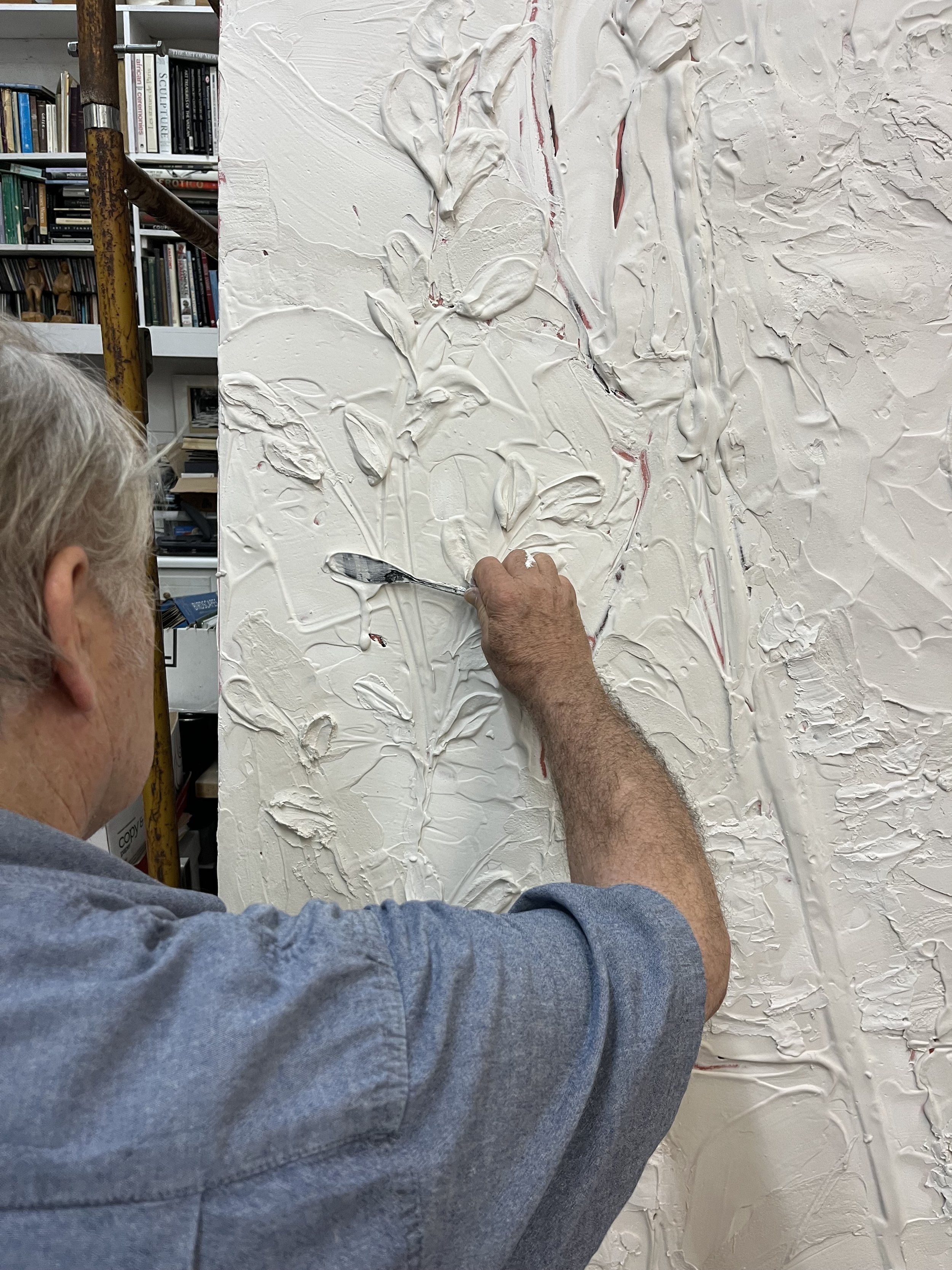 An artist applying thick white paint to a textured canvas with a palette knife, with a bookcase filled with books in the background.