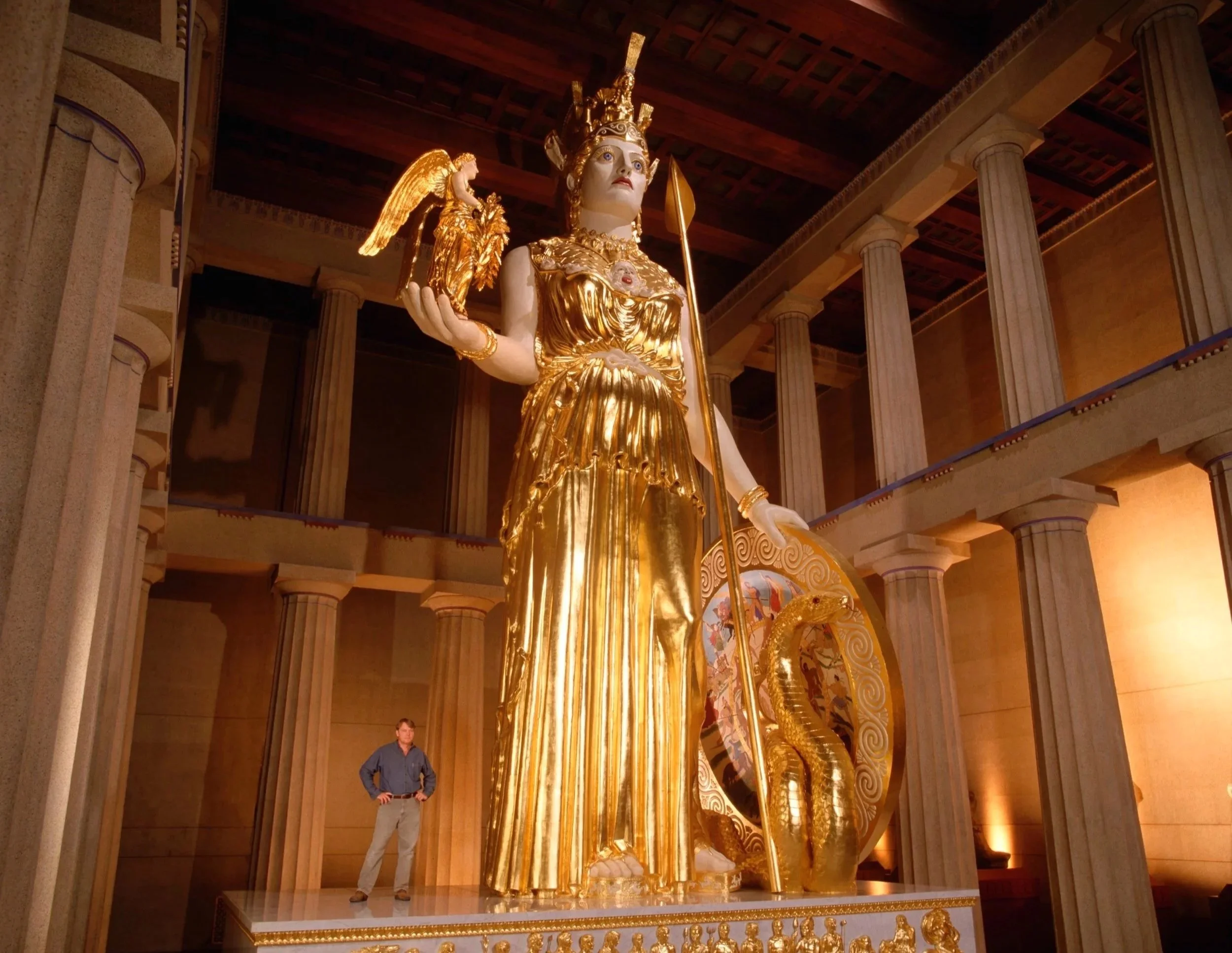 Grand golden statue of a regal woman with a crown, holding a staff, adorned with a necklace and a baby, in a large hall with tall columns and a wooden ceiling. A person stands near the statue, looking at it.