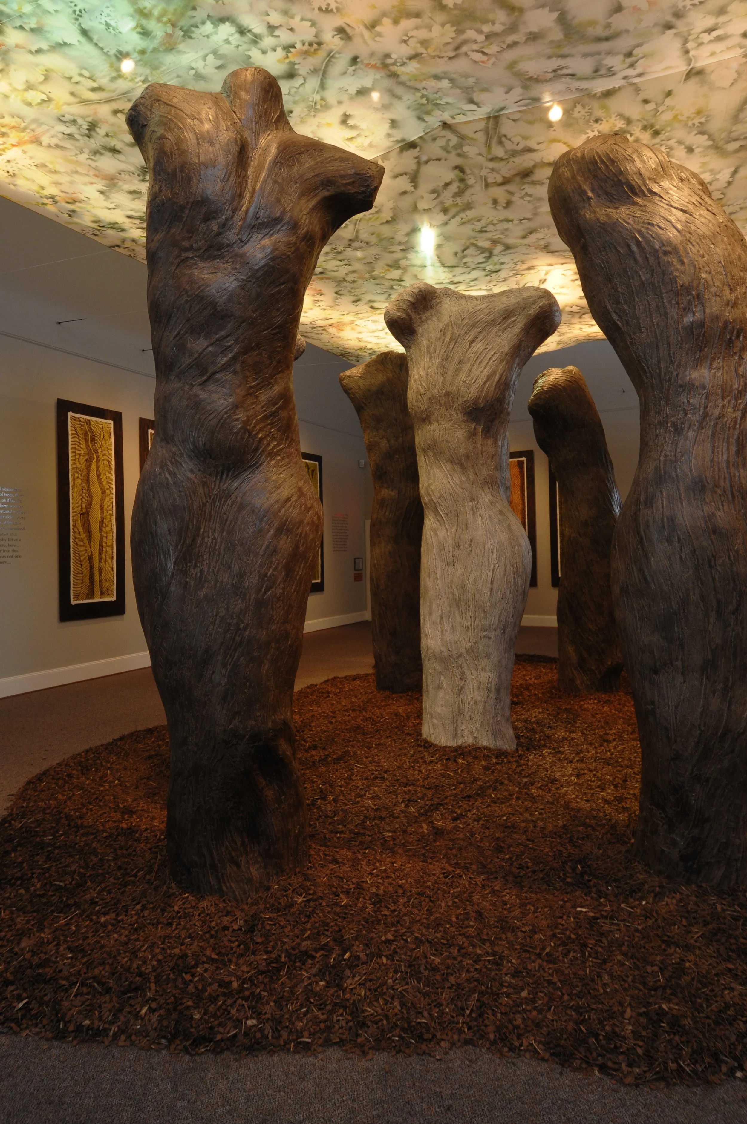 Four large, wooden, tree-trunk sculptures in an indoor art exhibit. The ceiling has a floral design, and framed artwork is on the wall in the background.