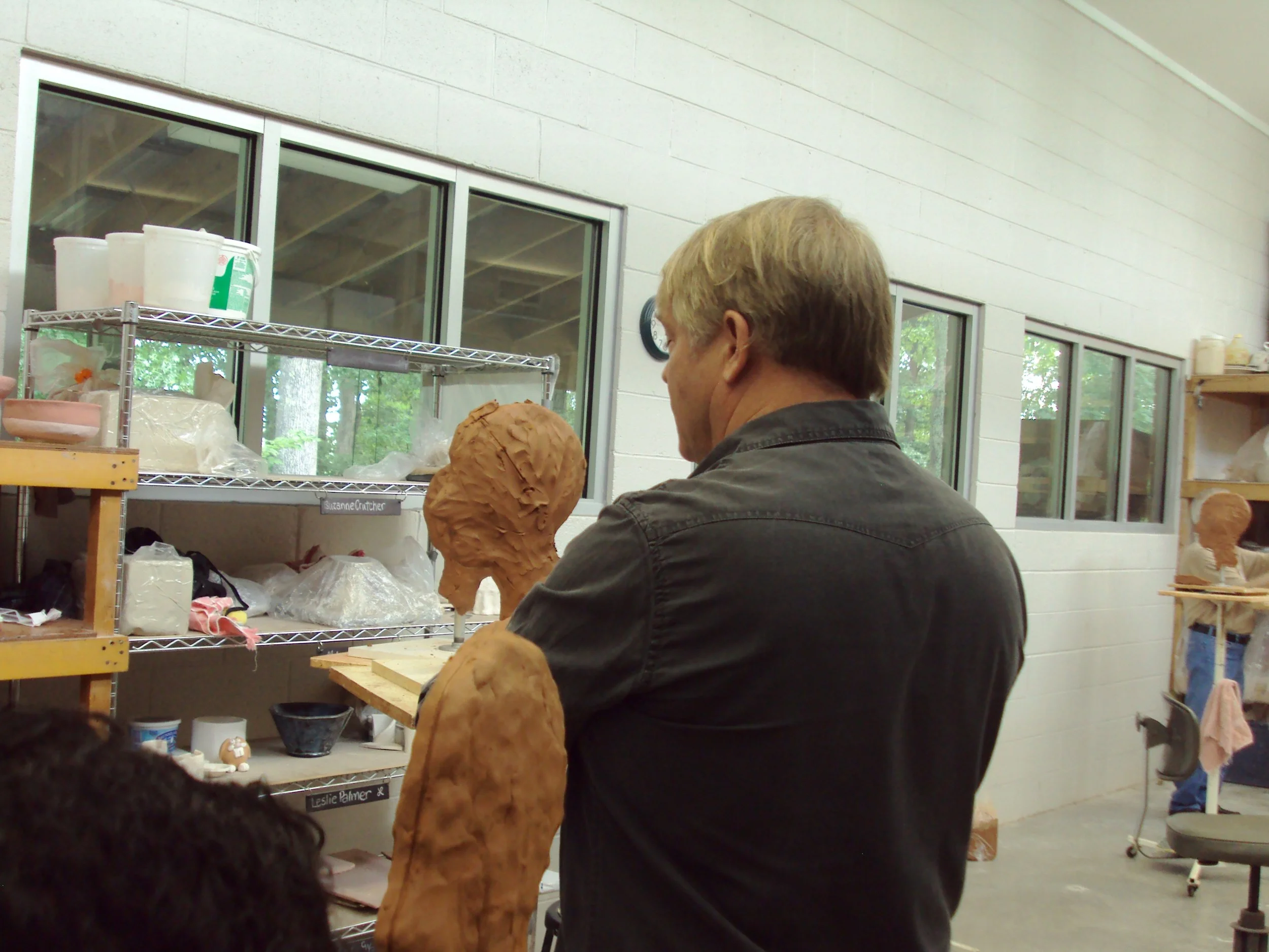 An artist works on a large clay bust in a ceramics studio, with shelves holding supplies and a window showing trees outside.
