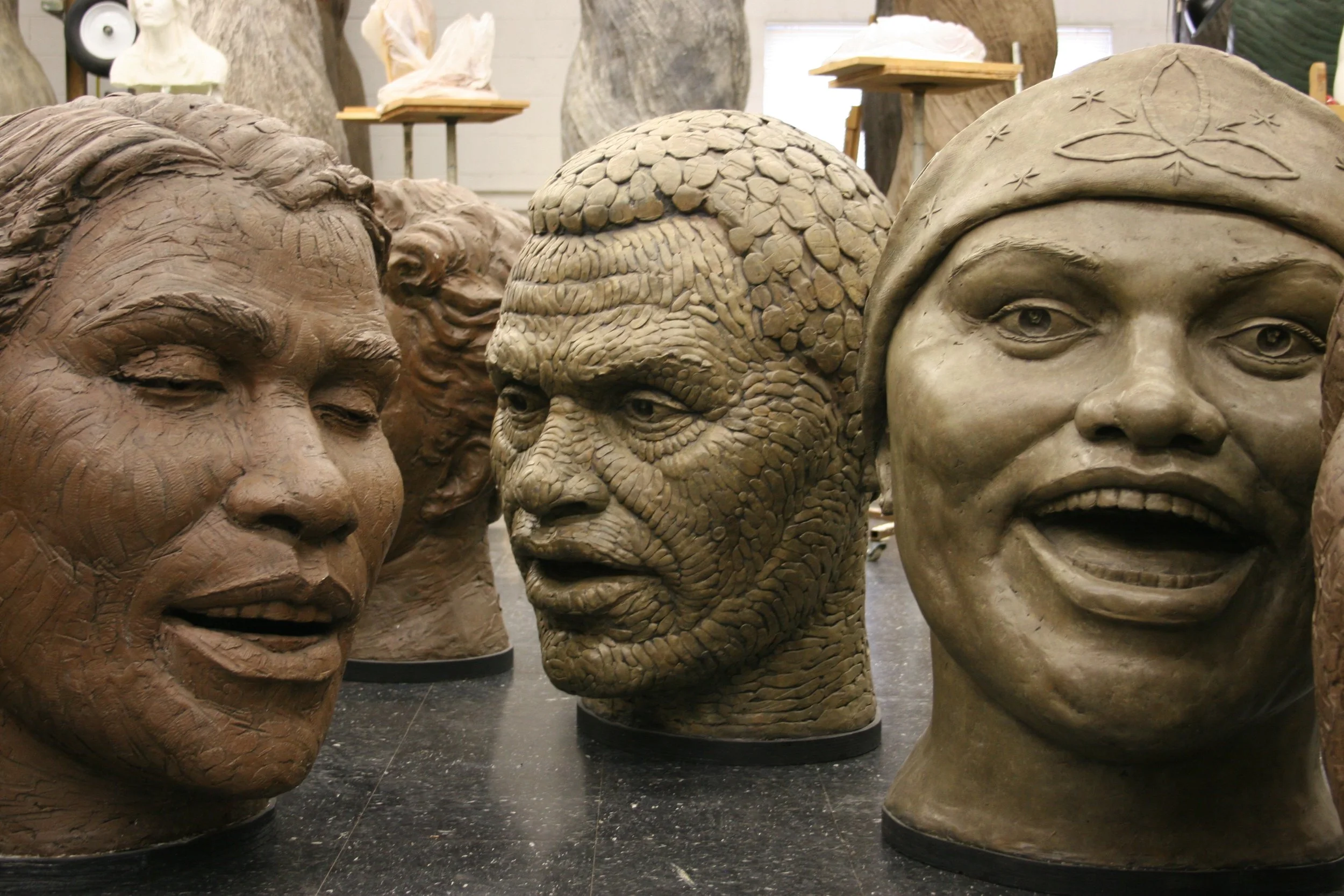 Several large sculptures of human heads with diverse facial features and hairstyles are displayed in an indoor art studio.  Cultural Heroes