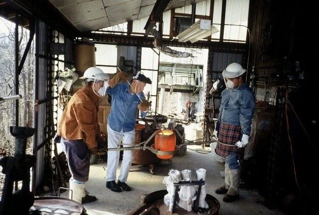 Blacksmiths working with heated metal inside a workshop, wearing safety gear including helmets and gloves.