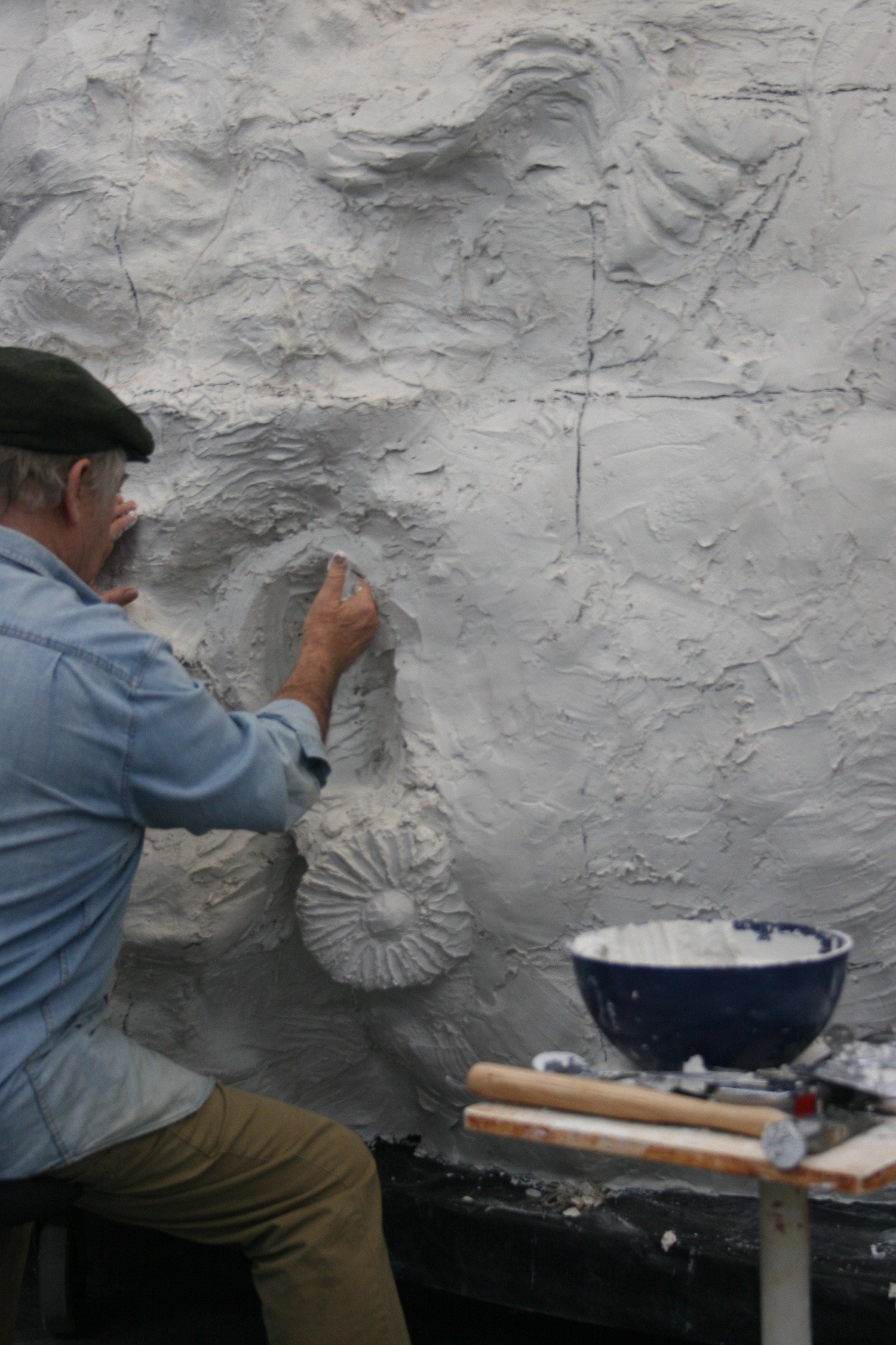 An artist is creating a large, detailed bas-relief sculpture on a wall, depicting a human face with closed eyes and wavy hair, in a studio setting.