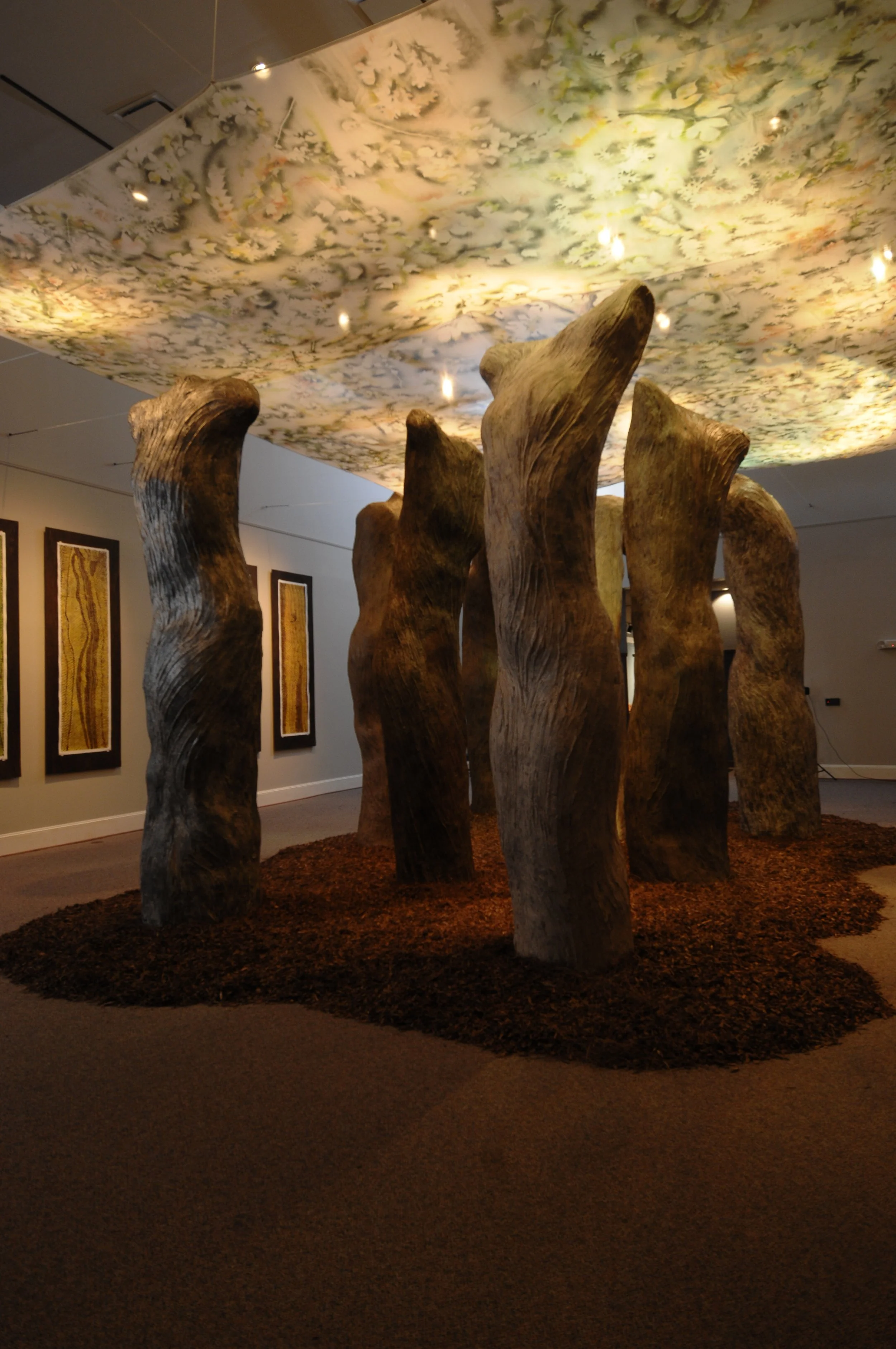 Art installation featuring tall, twisted wooden sculptures on a brown, textured surface, with a ceiling display of floral patterns and artwork on the walls.