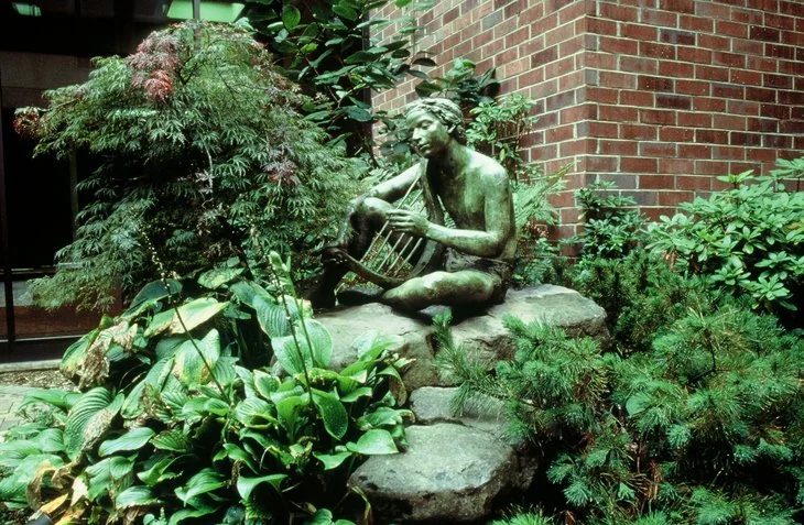 Bronze statue of a woman playing a stringed instrument, seated on rocks surrounded by lush green plants and bushes, with a brick wall in the background.