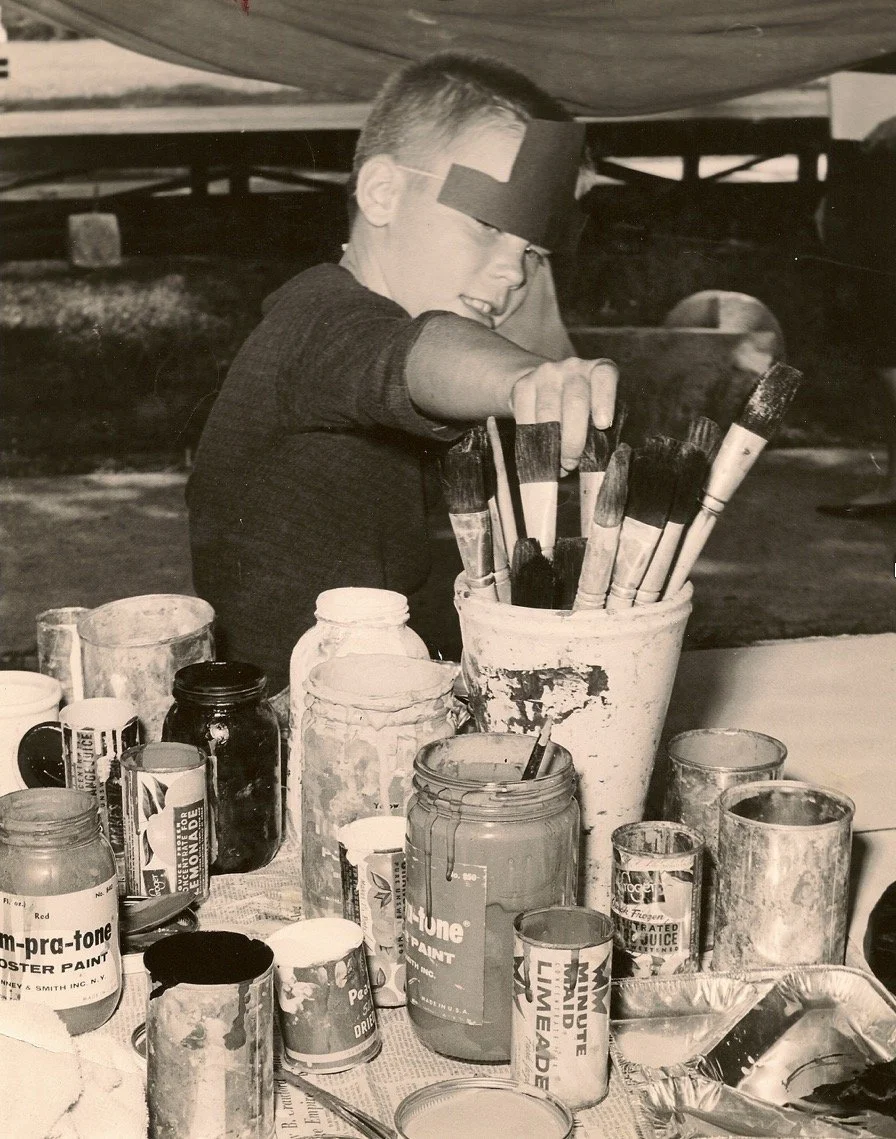 A young boy painting with brushes and paint jars on a table, wearing a black eye patch, in a workshop or art studio setting.
