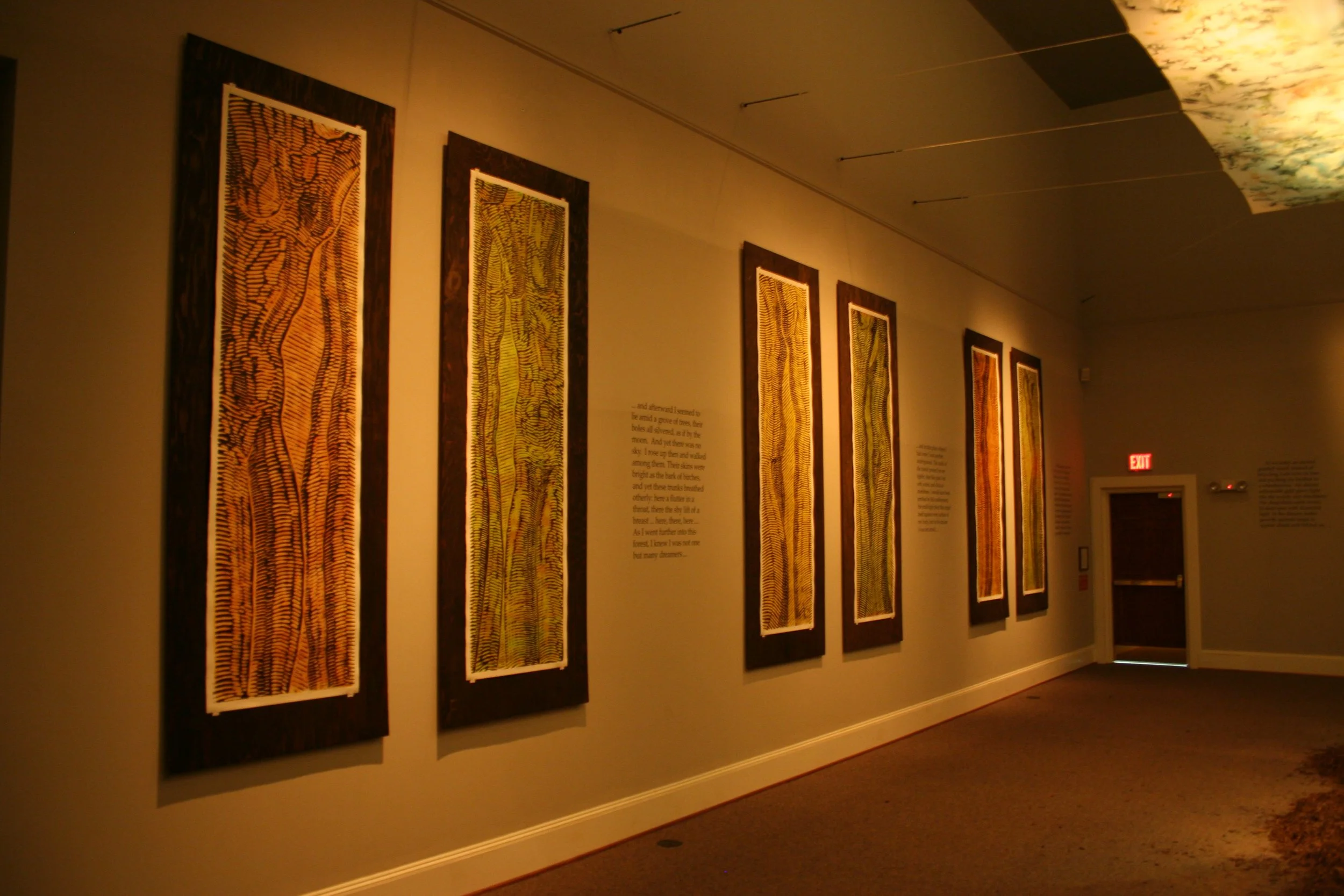 A hallway in an art gallery featuring vertically oriented artworks with intricate patterns, accompanied by text panels. Dream Forest Wood Block Prints.