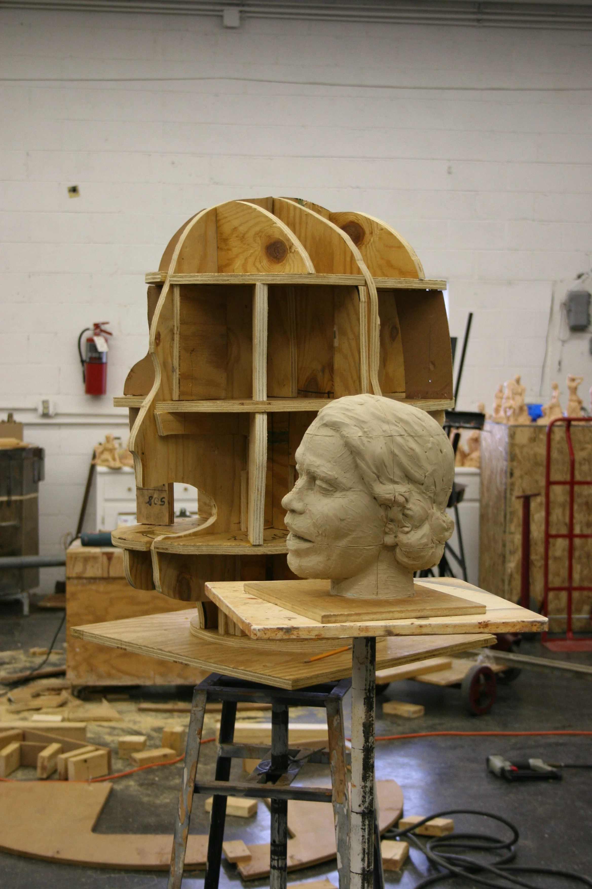 Art studio with unfinished wooden art pieces including a large head bust sculpture and a curved architectural model on standing tables, surrounded by wood scraps and tools.