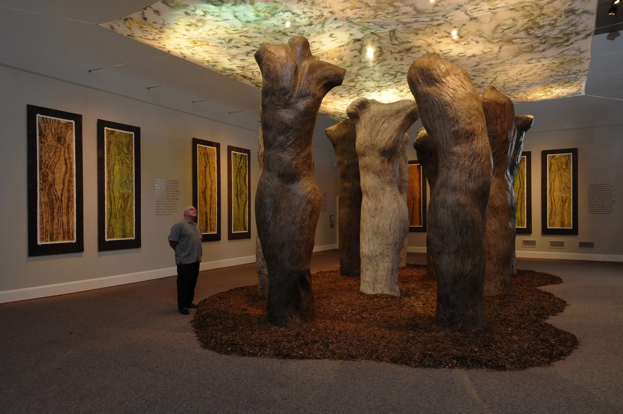 An art installation in a gallery featuring large, carved wooden sculptures resembling tree trunks arranged in a circle on a floor with a wood chip or bark surface. The ceiling has a floral or leaf pattern backlit with soft lighting. Two framed artwor