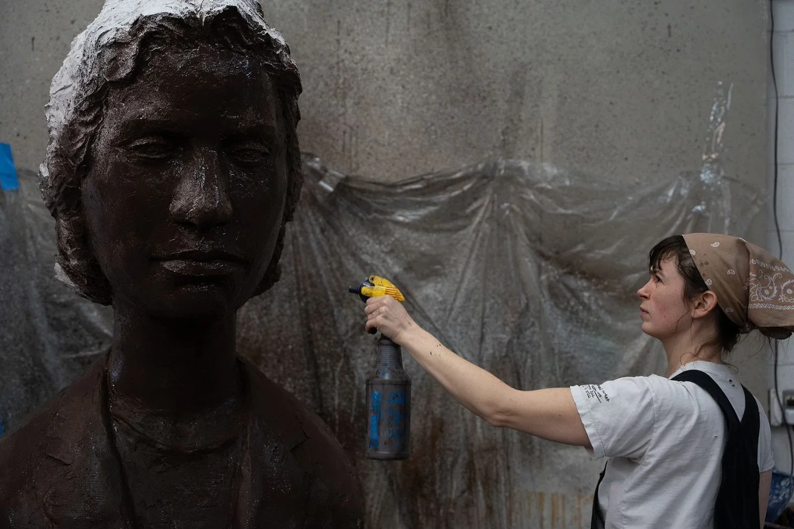 An artist is spray-painting a bronze statue of a person's head and shoulders against a gray wall covered with plastic.