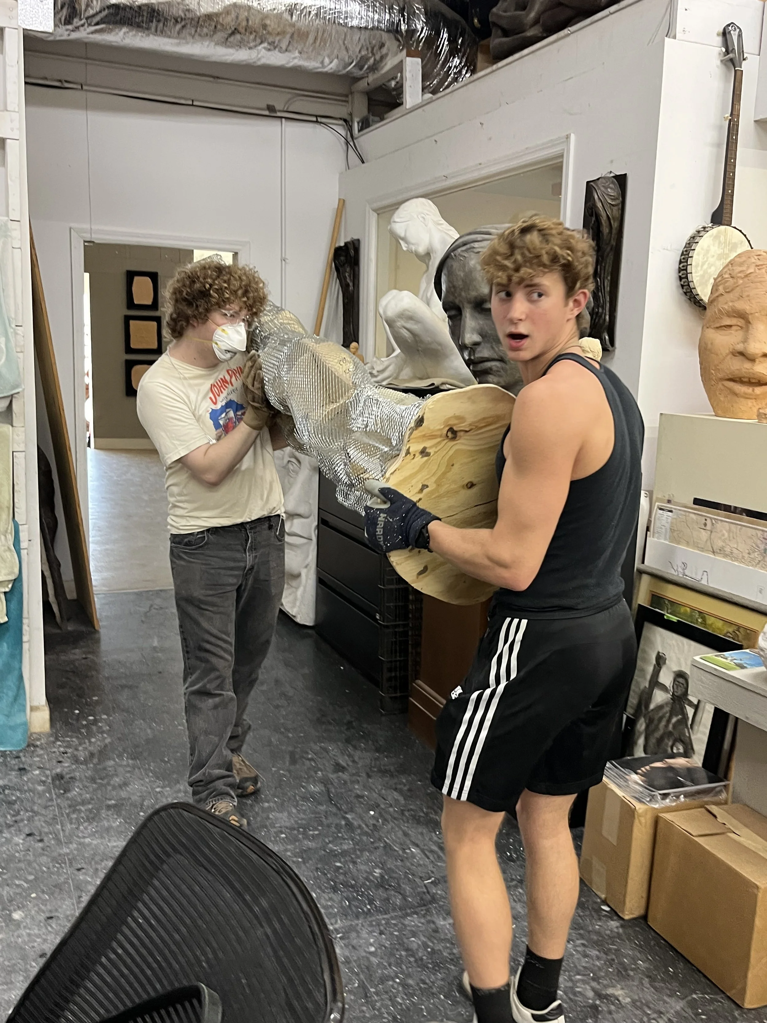 Two young men in a workshop or art studio working on a sculpture, with one holding a wooden base and the other attaching a metallic material to the sculpture, surrounded by various art pieces and supplies.