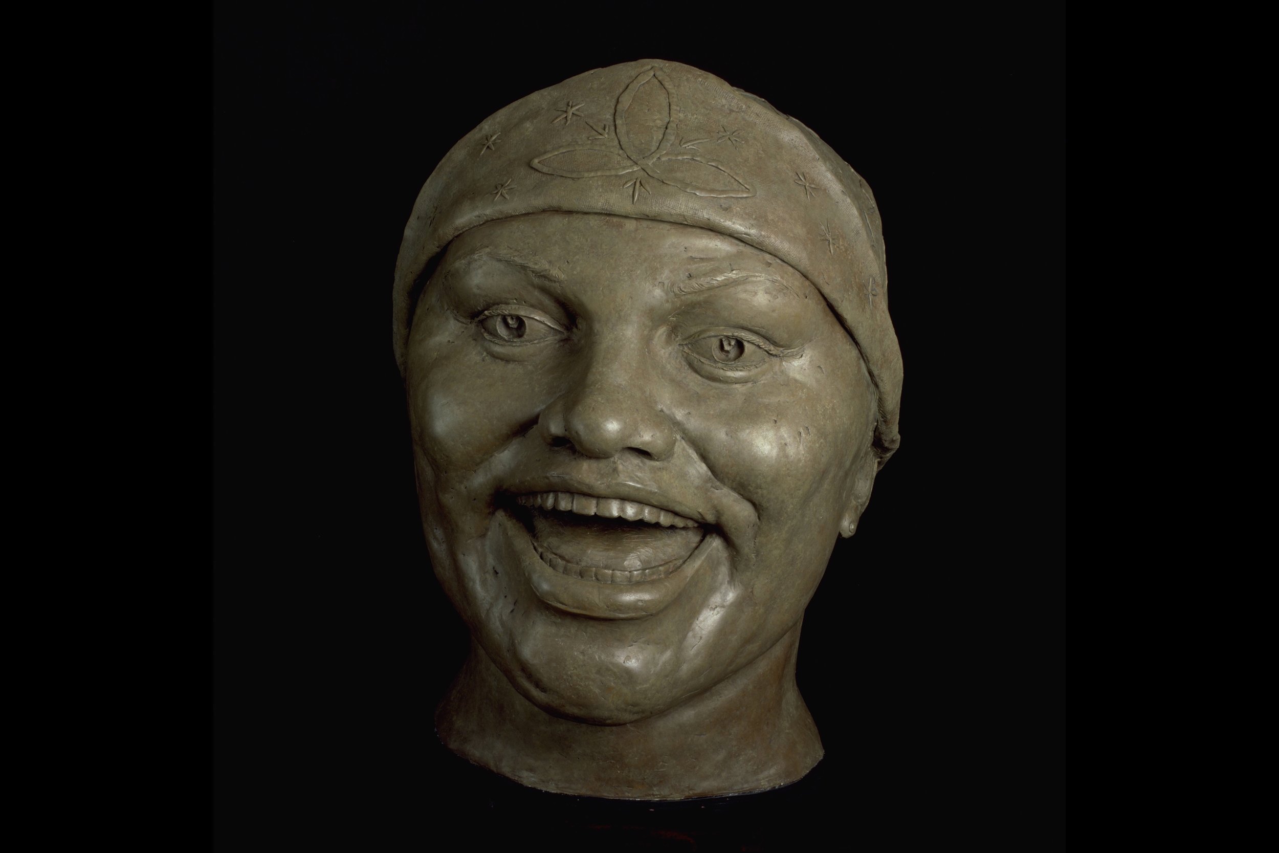 A sculpture of a smiling person's face with a head covering decorated with engraved leaves and stars, set against a black background.