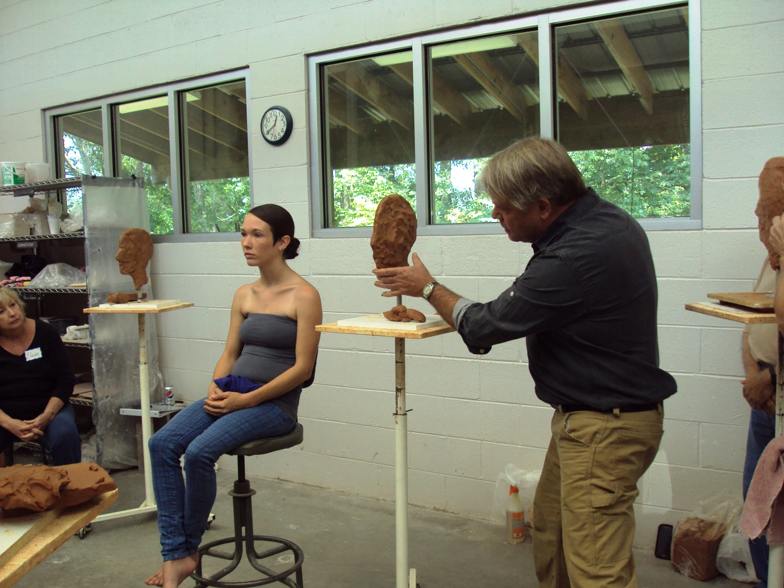A person with short brown hair is sculpting a large clay head on a stand in a room with large windows, while a woman with dark hair sitting nearby watches. A man with gray hair is examining a clay head on a stand, and another person with a clay head 