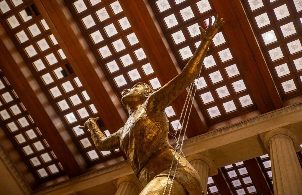 A large golden statue of a woman with outstretched arms, viewed from below, with a decorative ceiling and columns in the background.