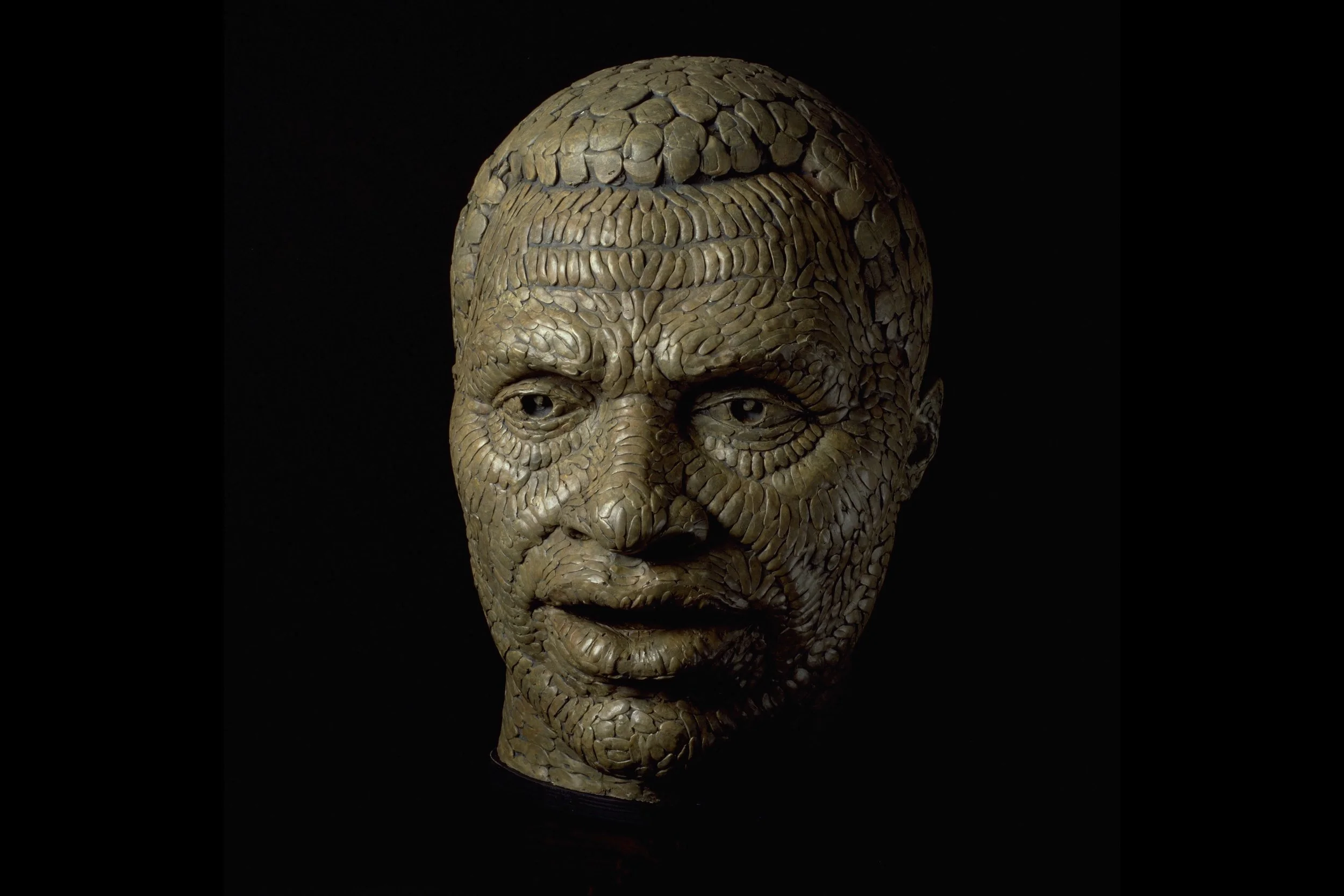 An artistic sculpture of a human face made from numerous small, textured, beige objects resembling seeds or stones, set against a black background.