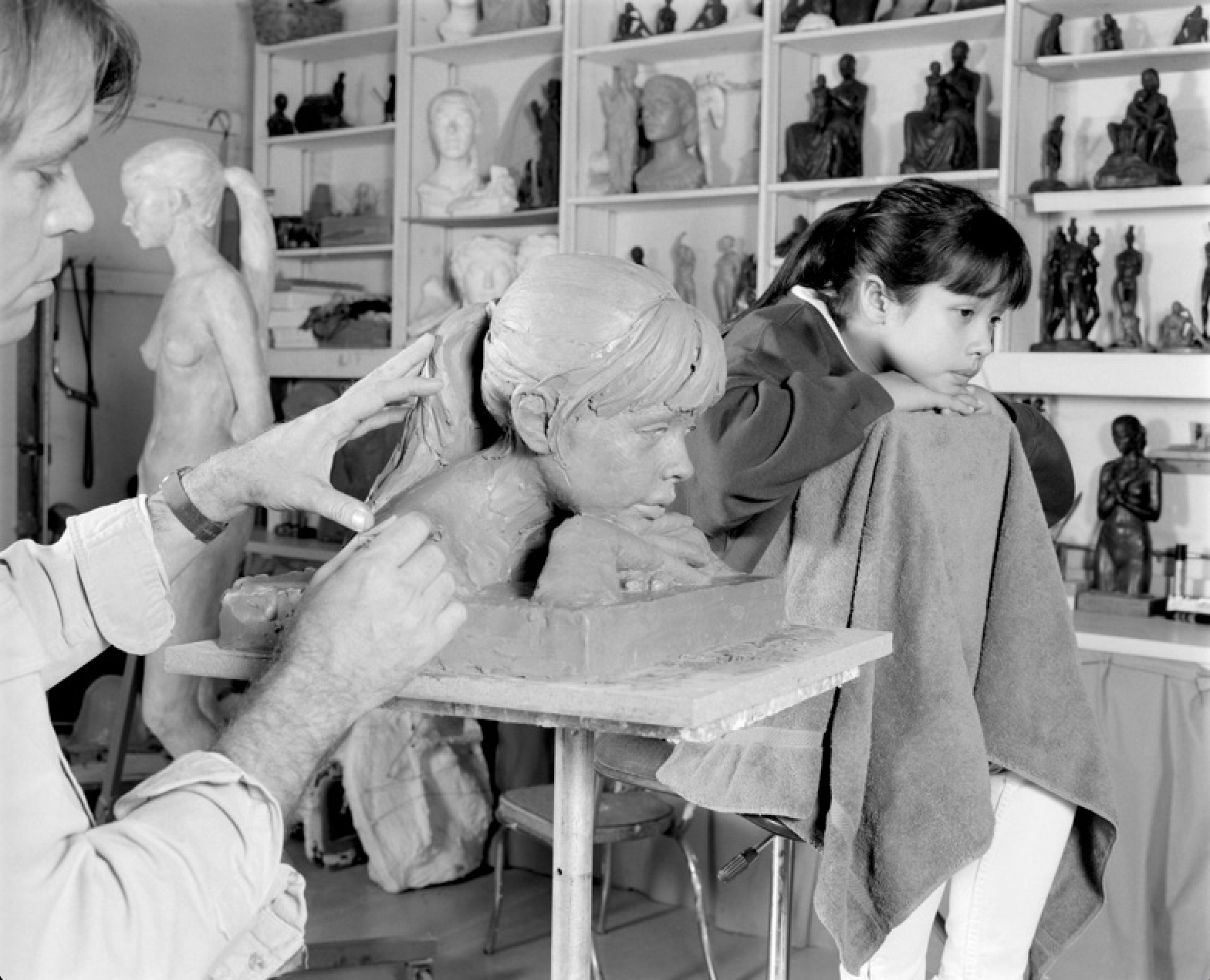 A person sculpting a clay bust of a woman with a headscarf, while a young girl observes. The room contains shelves with various sculptures and busts.