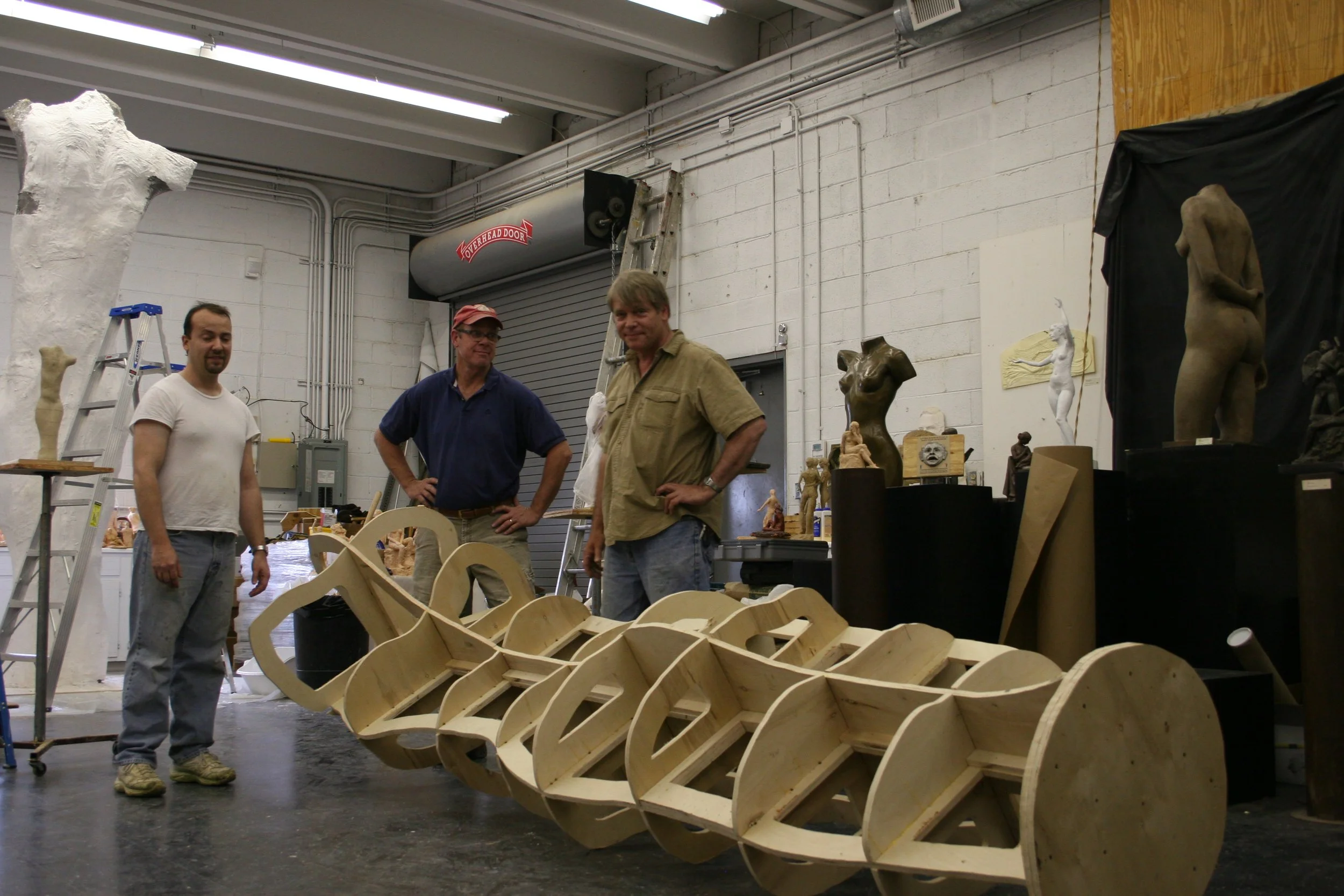 Three men standing inside a workshop with sculpture and woodworking projects; one man is wearing a white t-shirt, another a dark blue shirt and hat, and the third a tan shirt; large wooden sculpture framework and other sculptures are visible in the b