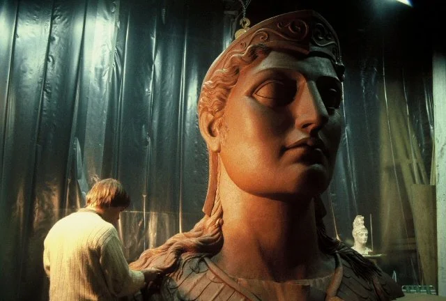 A person working on a large statue of a woman with classical features, made of clay, in a workshop or studio setting. Athena Parthenos in Parthenon Museum Nashville. 