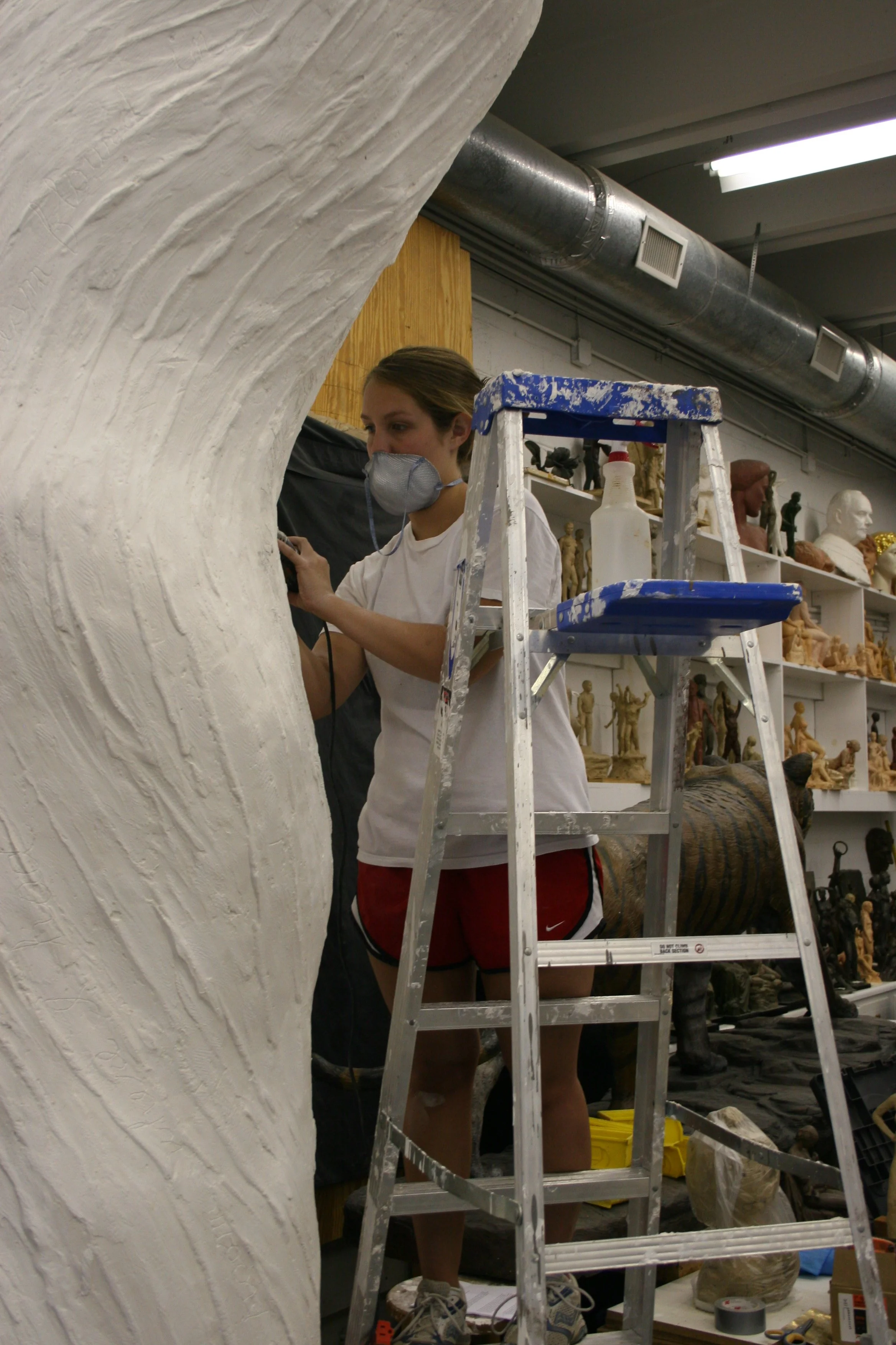 A person working on a sculpture, wearing a mask, surrounded by art supplies and sculptures in a studio.