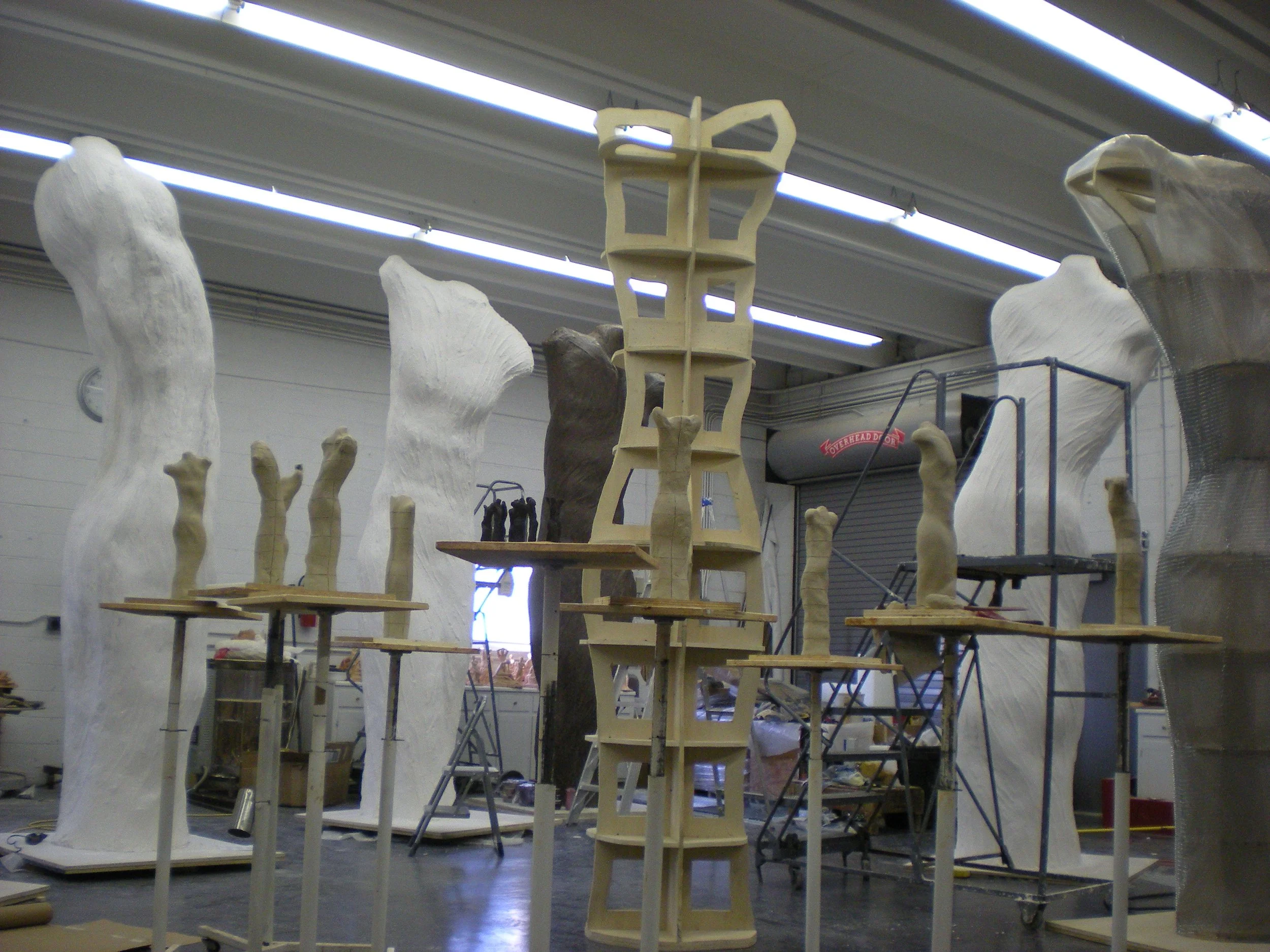 Workshop with various sculptures and foam structures in progress, including a tall, narrow armature sculpture in the center and large, curved white forms around it.