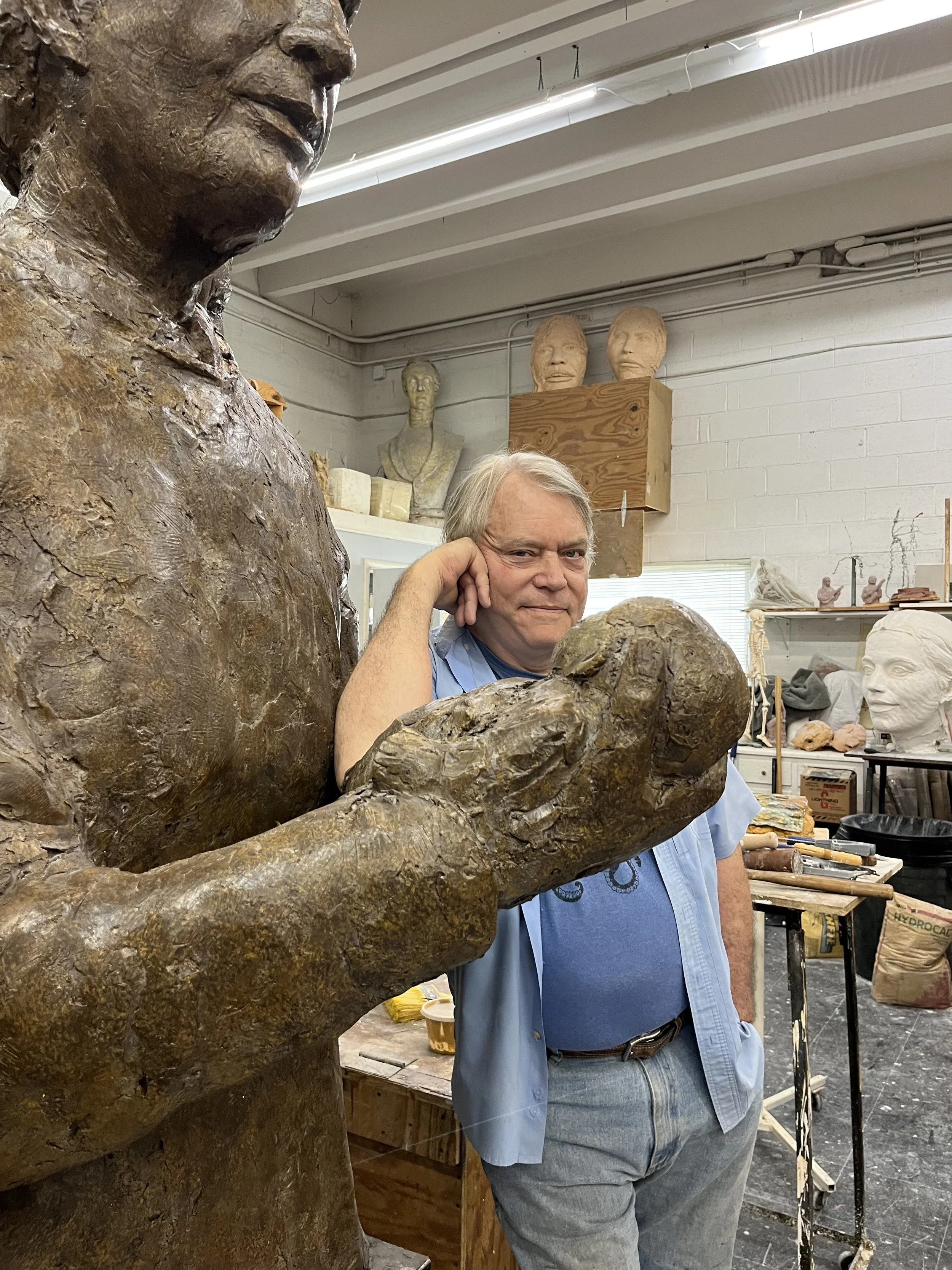 A man standing in an art studio or sculpting workshop, surrounded by sculptures and art supplies, resting his head on his hand while posing next to a large bronze sculpture of a woman with an expression of contentment.