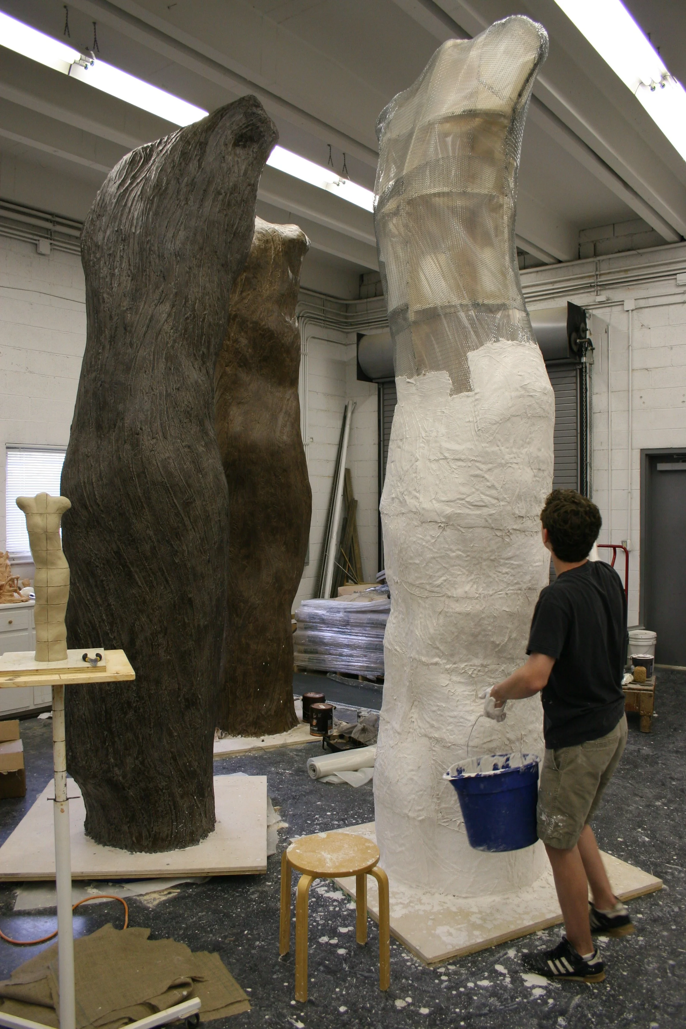 A person in a workshop adding plaster to a tall sculpture of a human torso, with other large sculptures, tools, and supplies around.