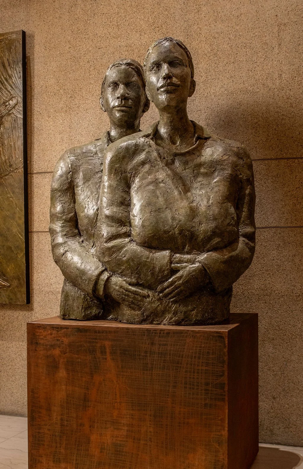 Bronze sculpture of two women standing close together against a beige wall.