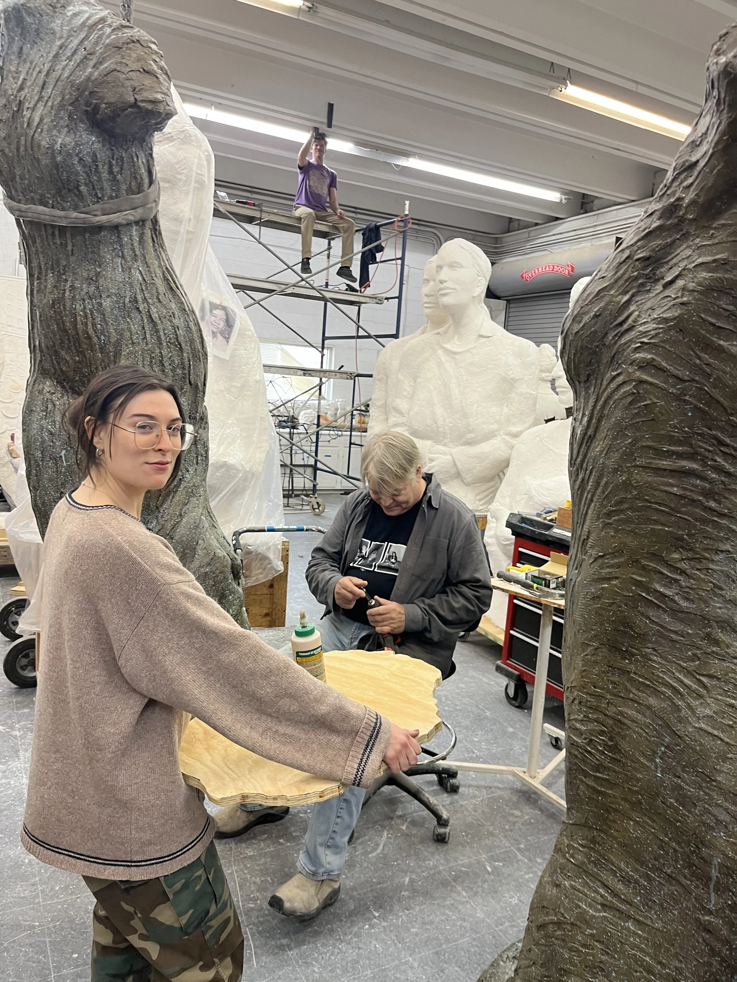 People working on a large sculpture in a workshop, with scaffolding and various tools around