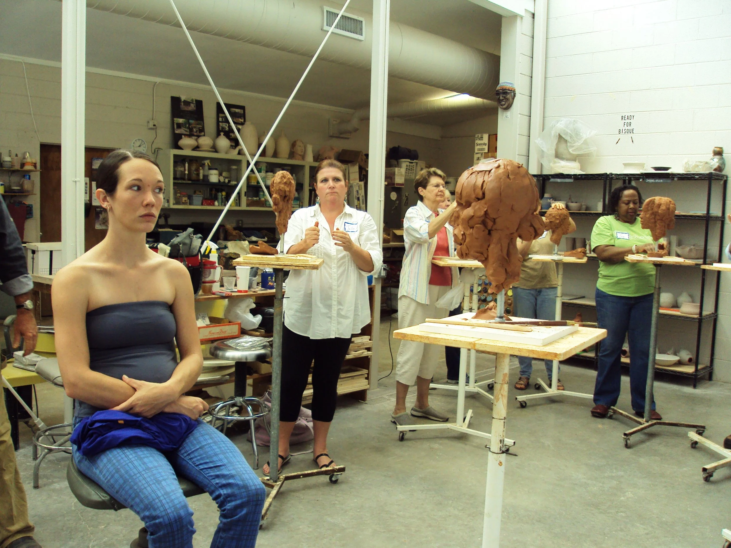 A group of women working on clay sculptures in an art studio. One woman is seated in the foreground, and others stand in the background shaping sculptures.