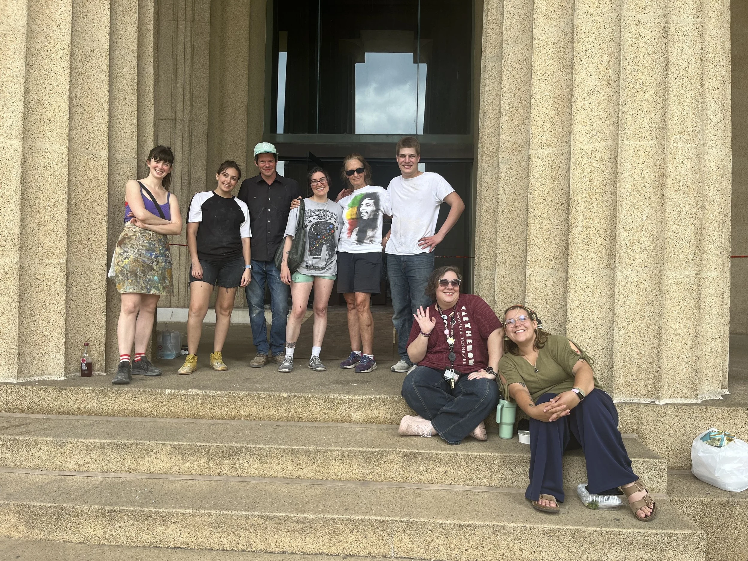 A group of nine people pose on the steps of a large building with columns and a glass entrance. Some are standing, and some are sitting, smiling and making various gestures. They are dressed casually, and some have drinks and personal items with them