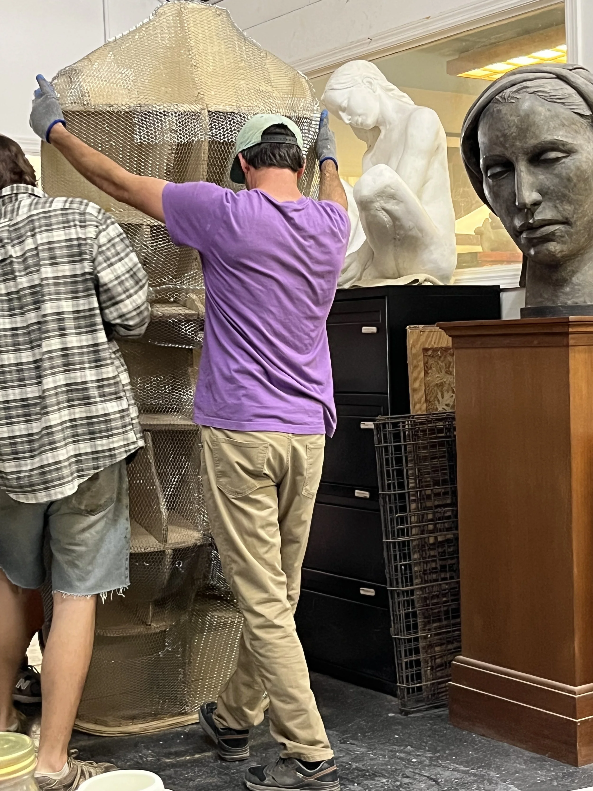 Two people working on a sculpture, one with a plaid shirt and another with a purple t-shirt, surrounded by sculptures and art supplies in a studio.