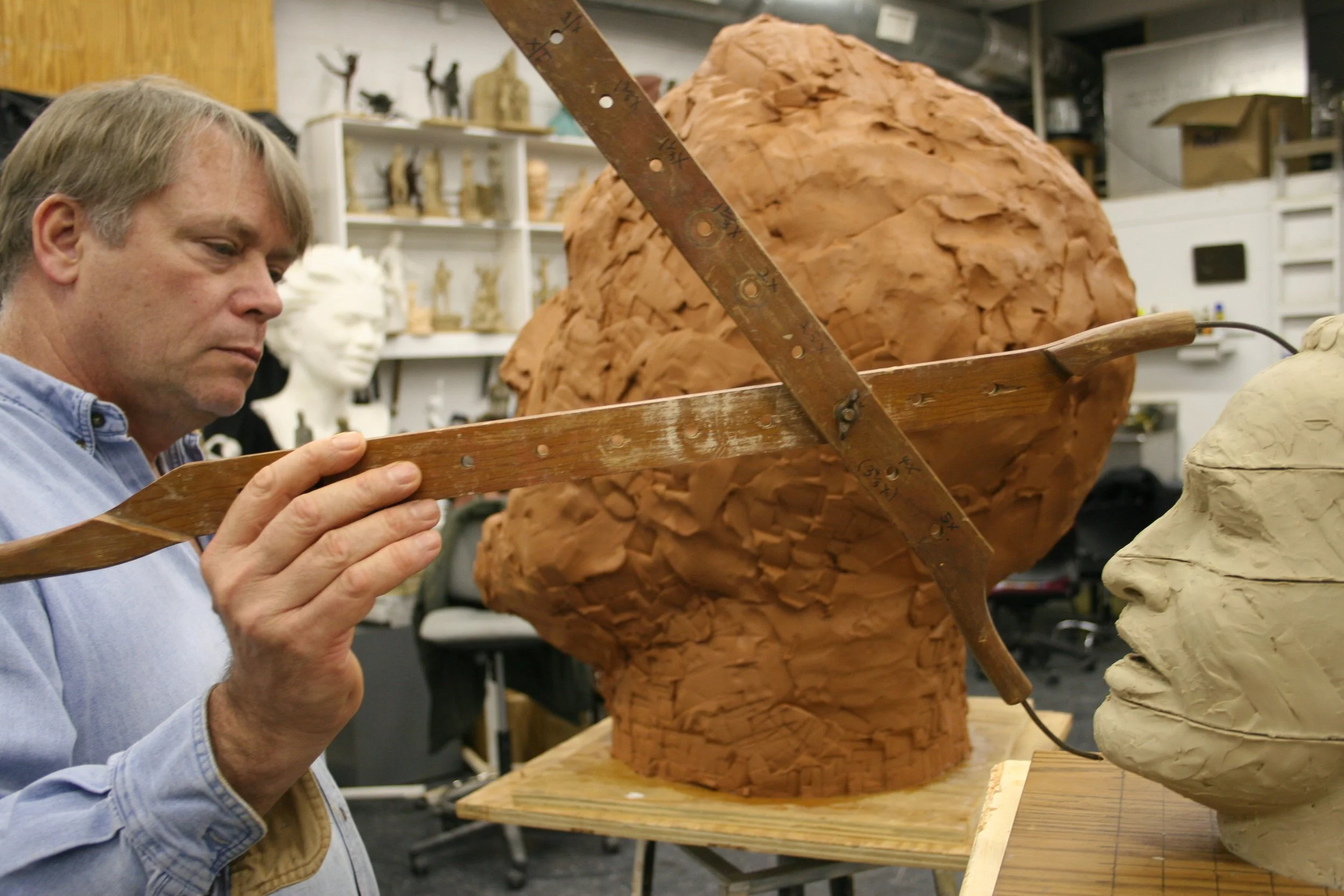 A man working on a wooden sculpture of a human head, using a measuring tool in a workshop filled with other sculptures and art pieces.