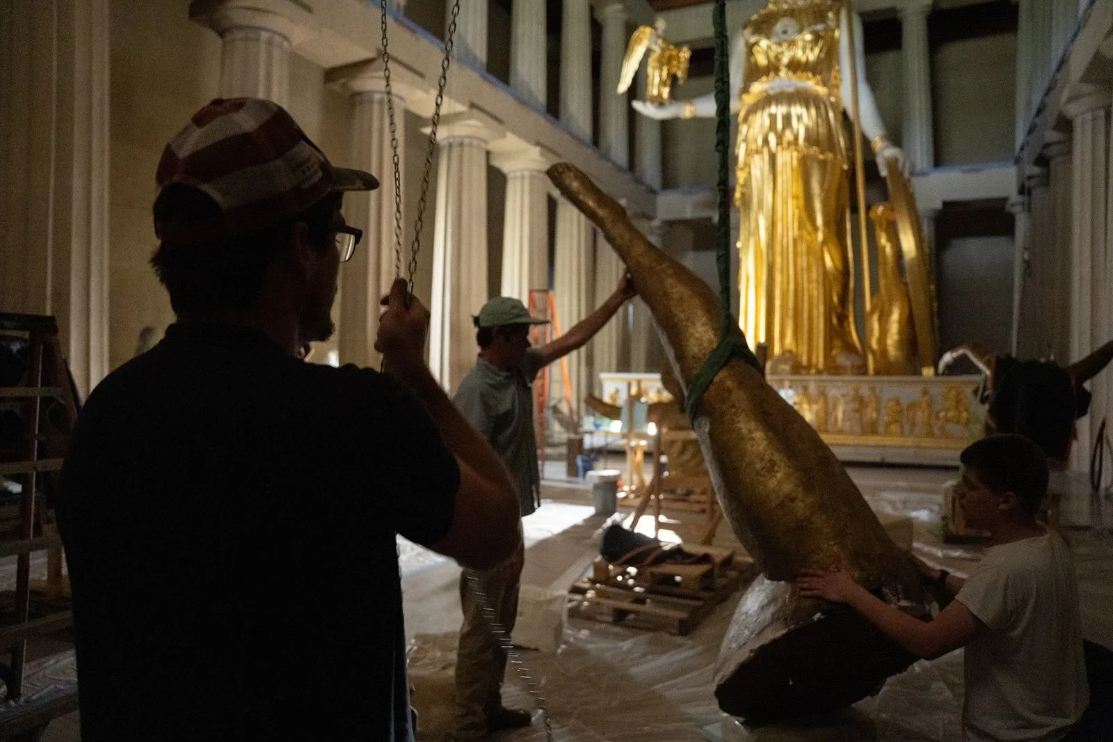 People working on lifting a large, golden sculpture inside a grand hall with tall columns and ornate decor.