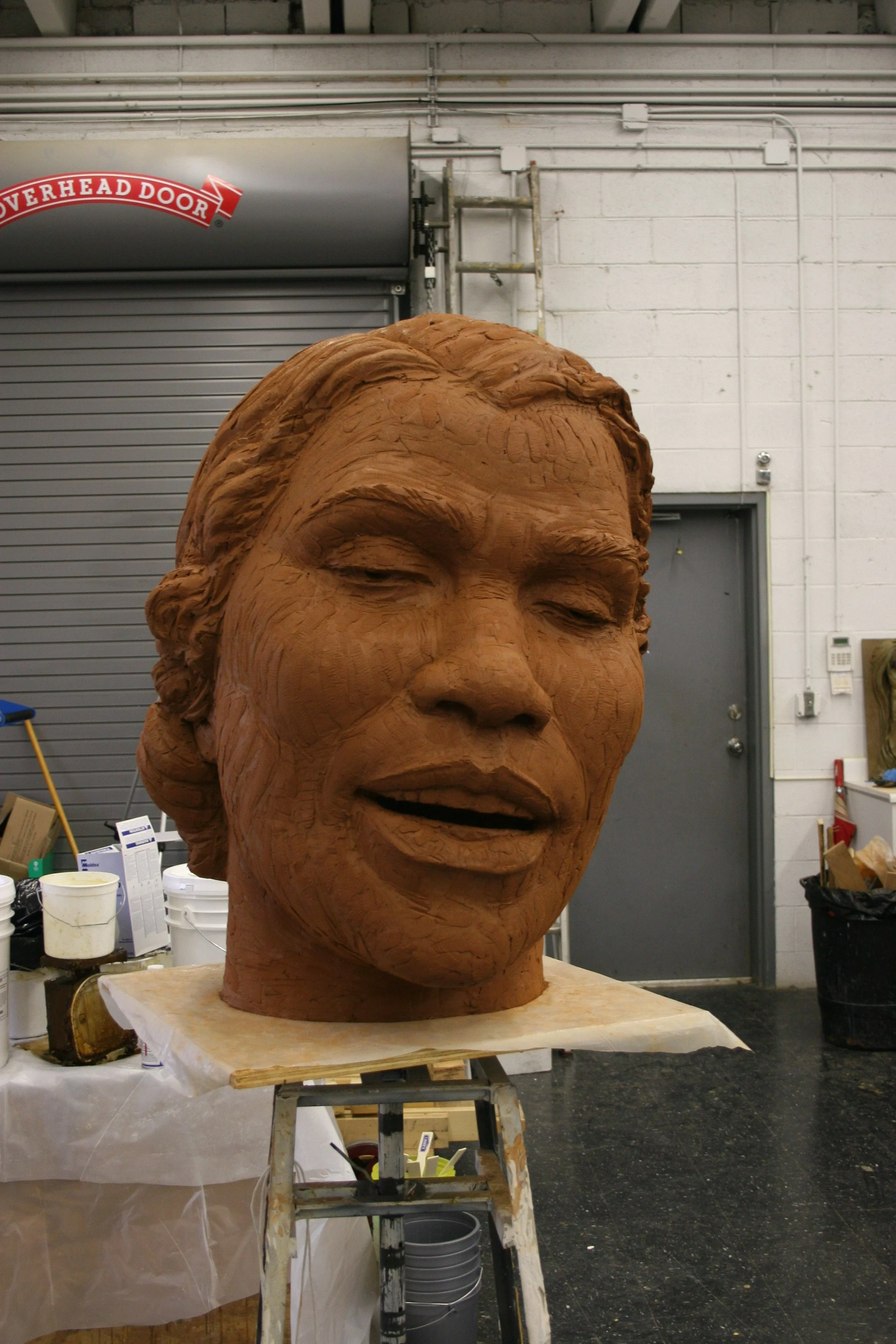 A large clay sculpture of a woman's head with closed eyes and a calm expression is in a workshop. The background includes a metal door, white brick wall, and various tools and supplies.